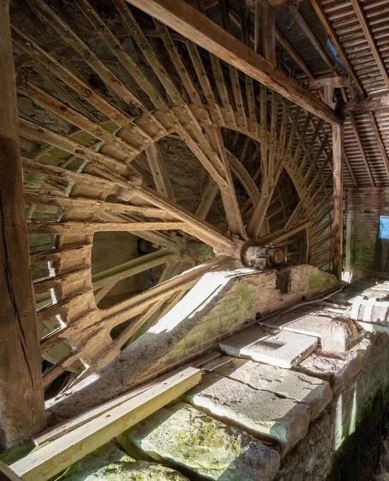 An old wooden watermill wheel inside a rustic building, with stone and wooden structures around it.