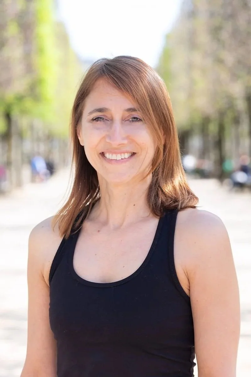 A woman with shoulder-length brown hair smiling outdoors on a sunny day, wearing a black tank top.
