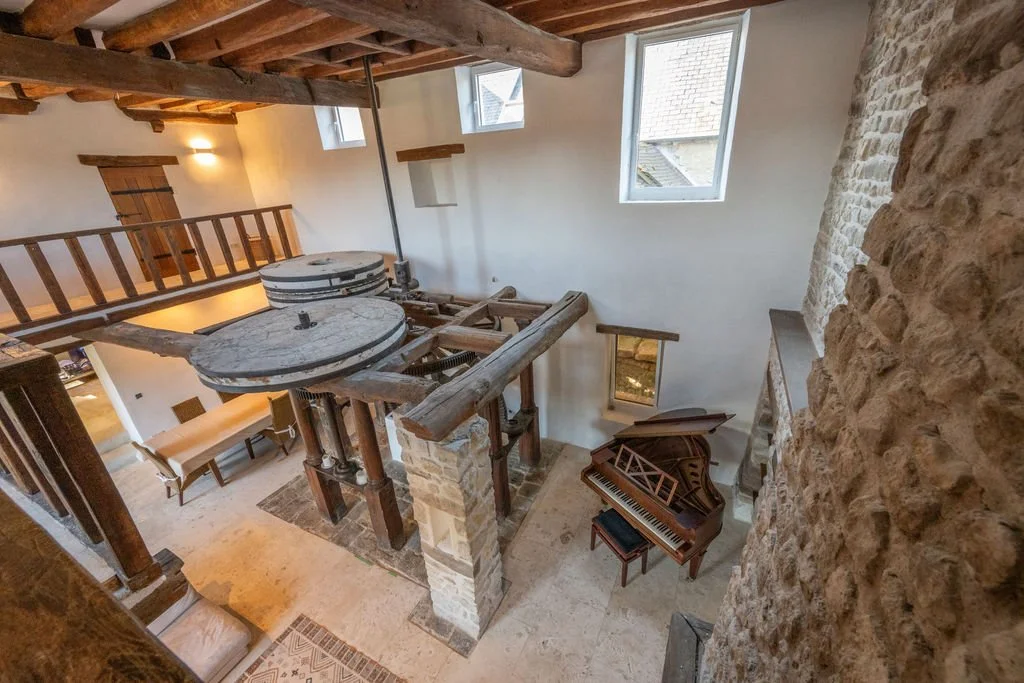 A rustic interior featuring a stone wall, wooden beams on the ceiling, a small staircase with a wooden railing, a grand piano, and three windows allowing natural light in.
