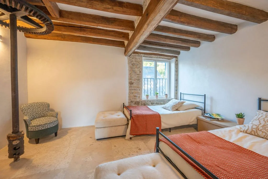 Two beds with metal frames, a small wooden nightstand with books, a potted plant, a window with a view outside, an armchair with a patterned fabric, a beige ottoman, exposed wooden ceiling beams, stone window frame, and beige tile flooring.