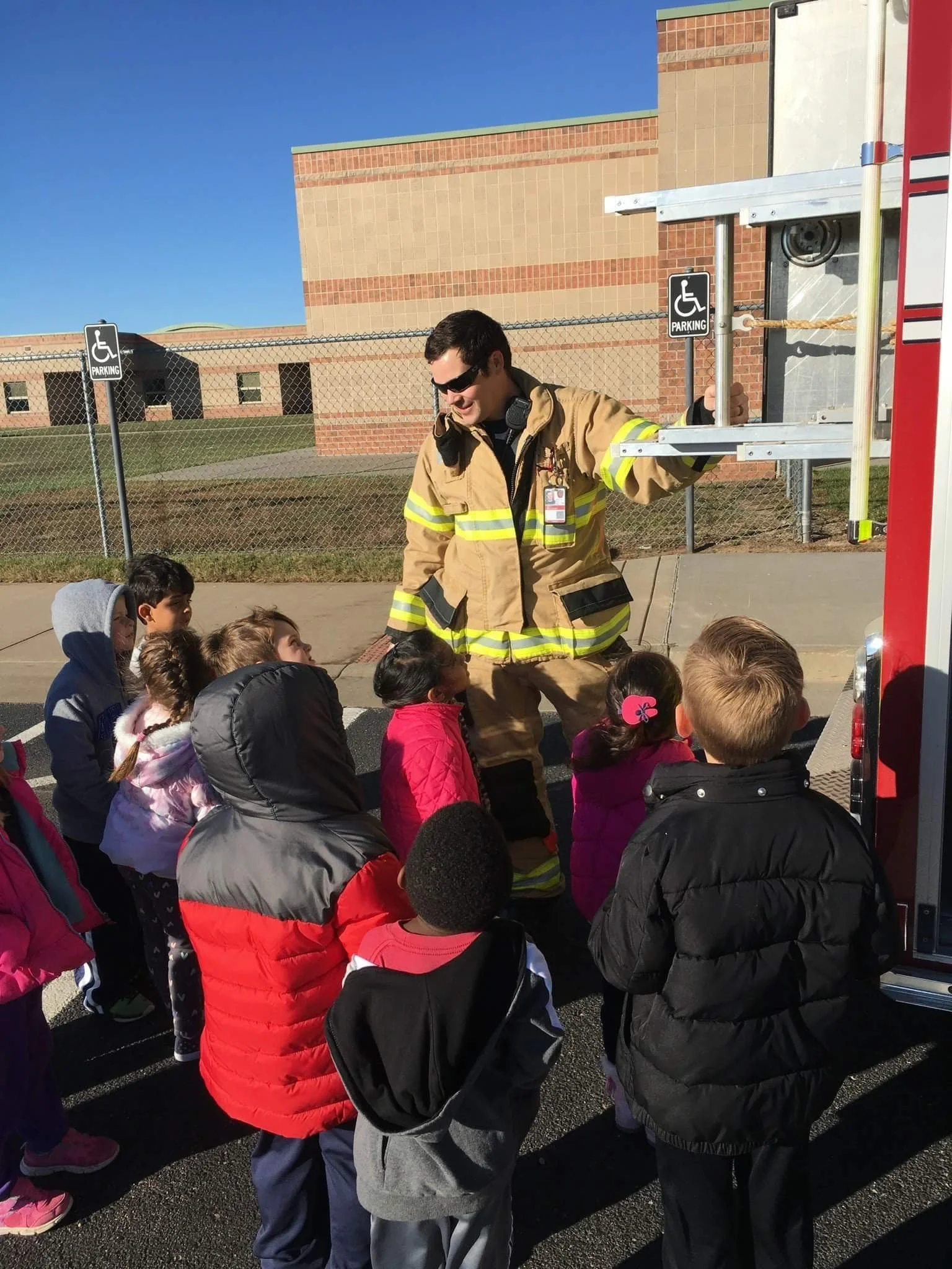 Owner Firefighter Joey Donaldson showing a group of children the inside of a fire truck outdoors on a sunny day.