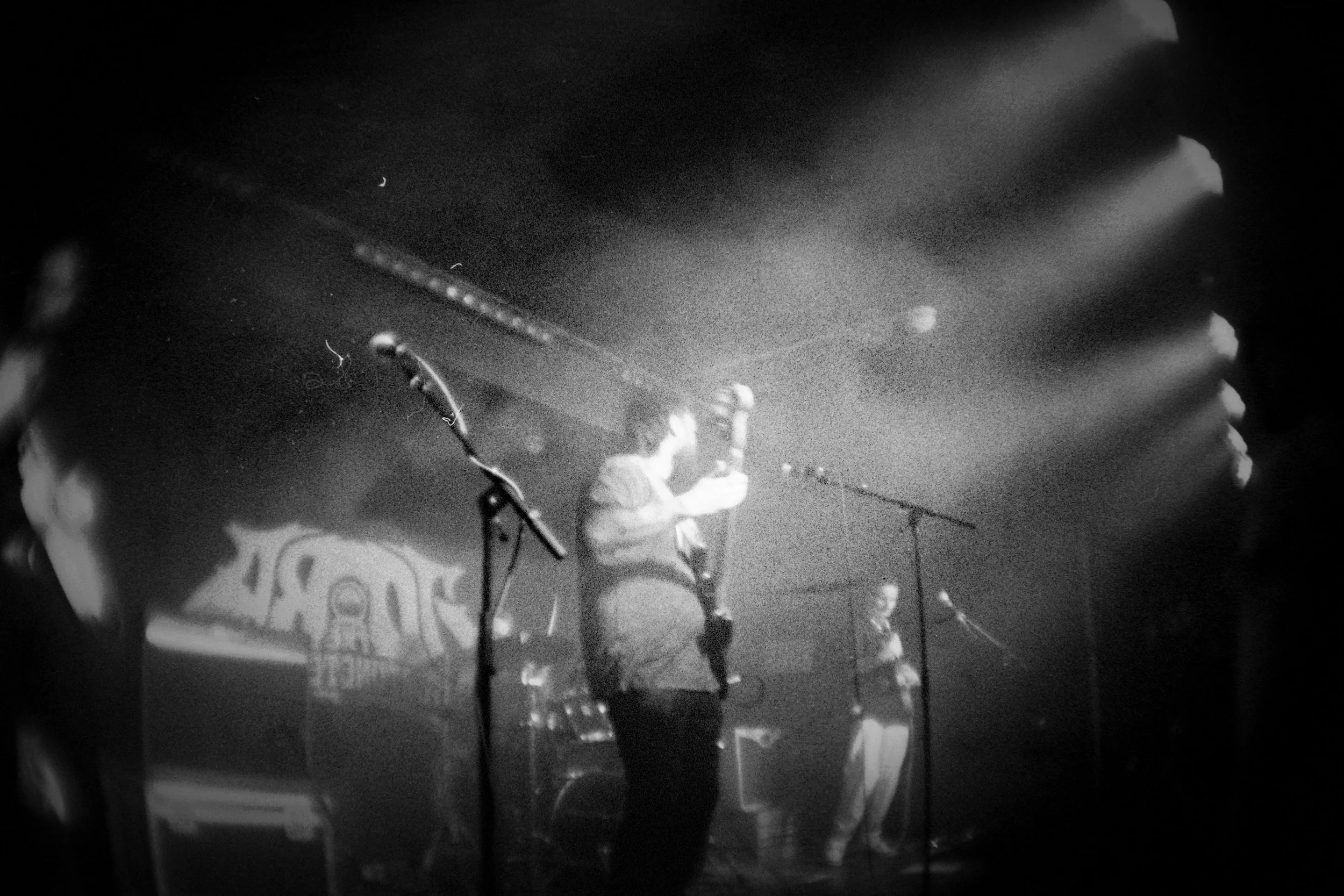 Black and white photo of two musicians performing on stage, with microphone stands and a drum set in the background.