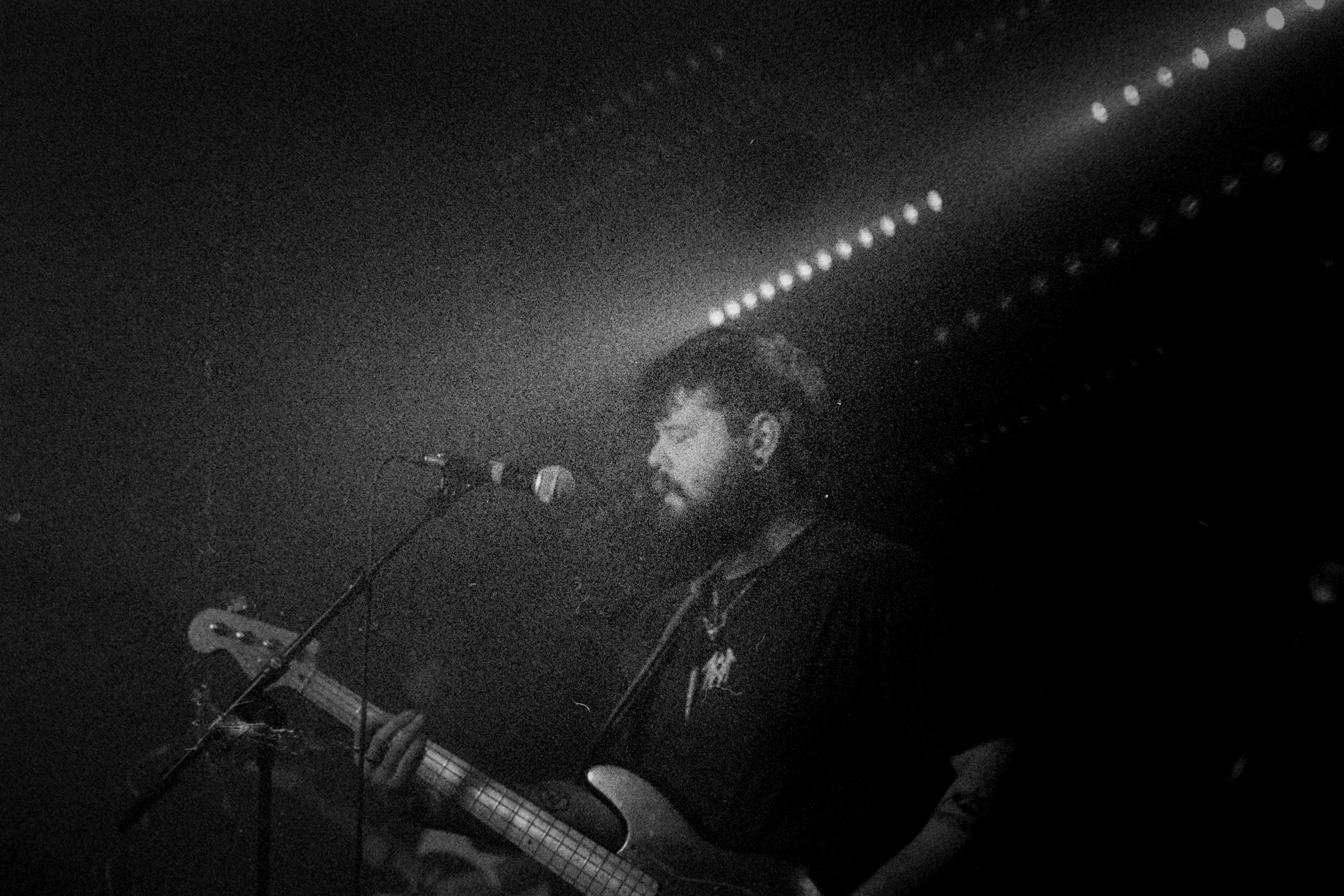 A man singing into a microphone while playing a guitar in a dark, illuminated setting with string lights overhead.