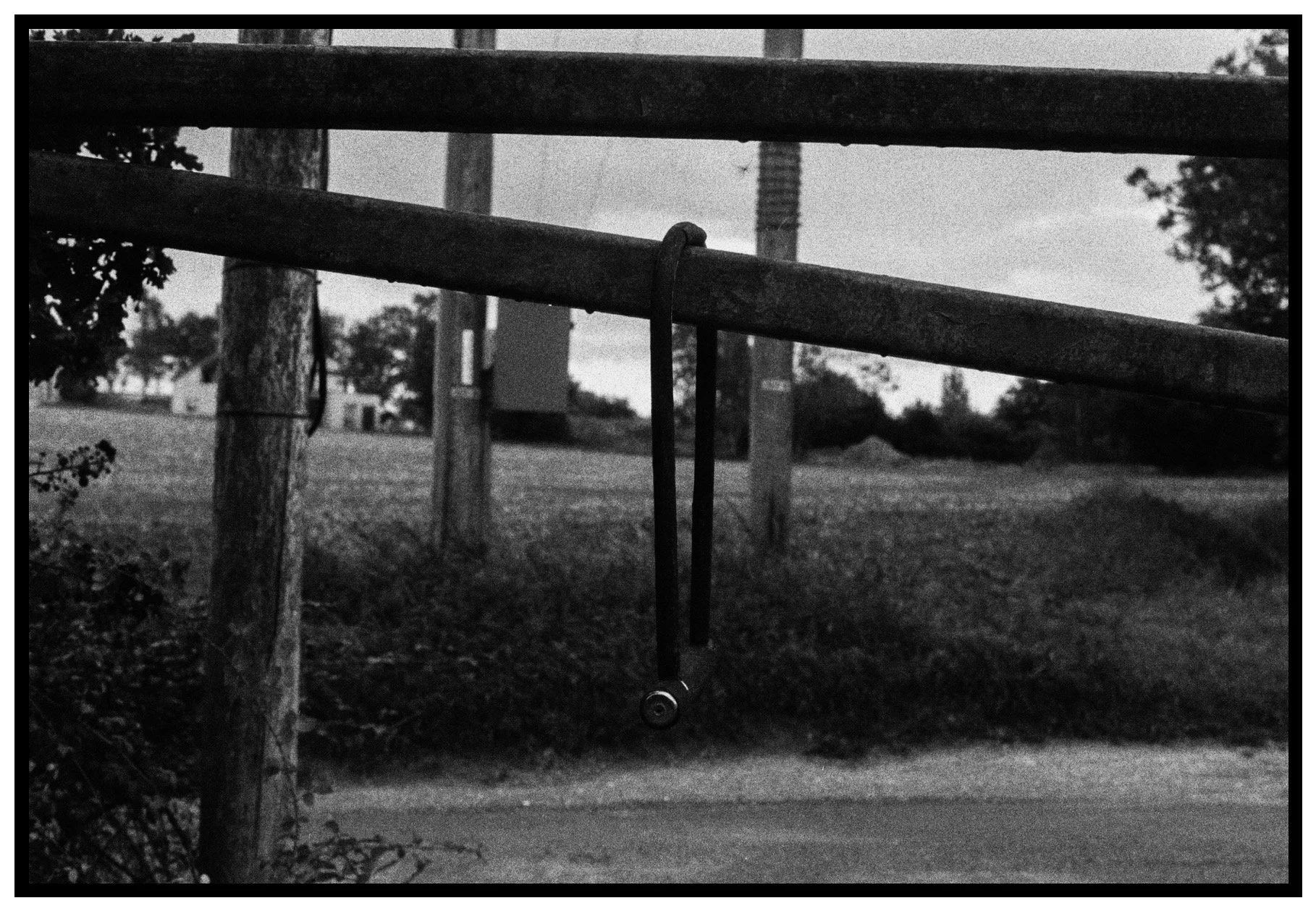 A black and white photo of a fidget spinner hanging from a horizontal wooden bar. There are trees in the background and a cloudy sky.