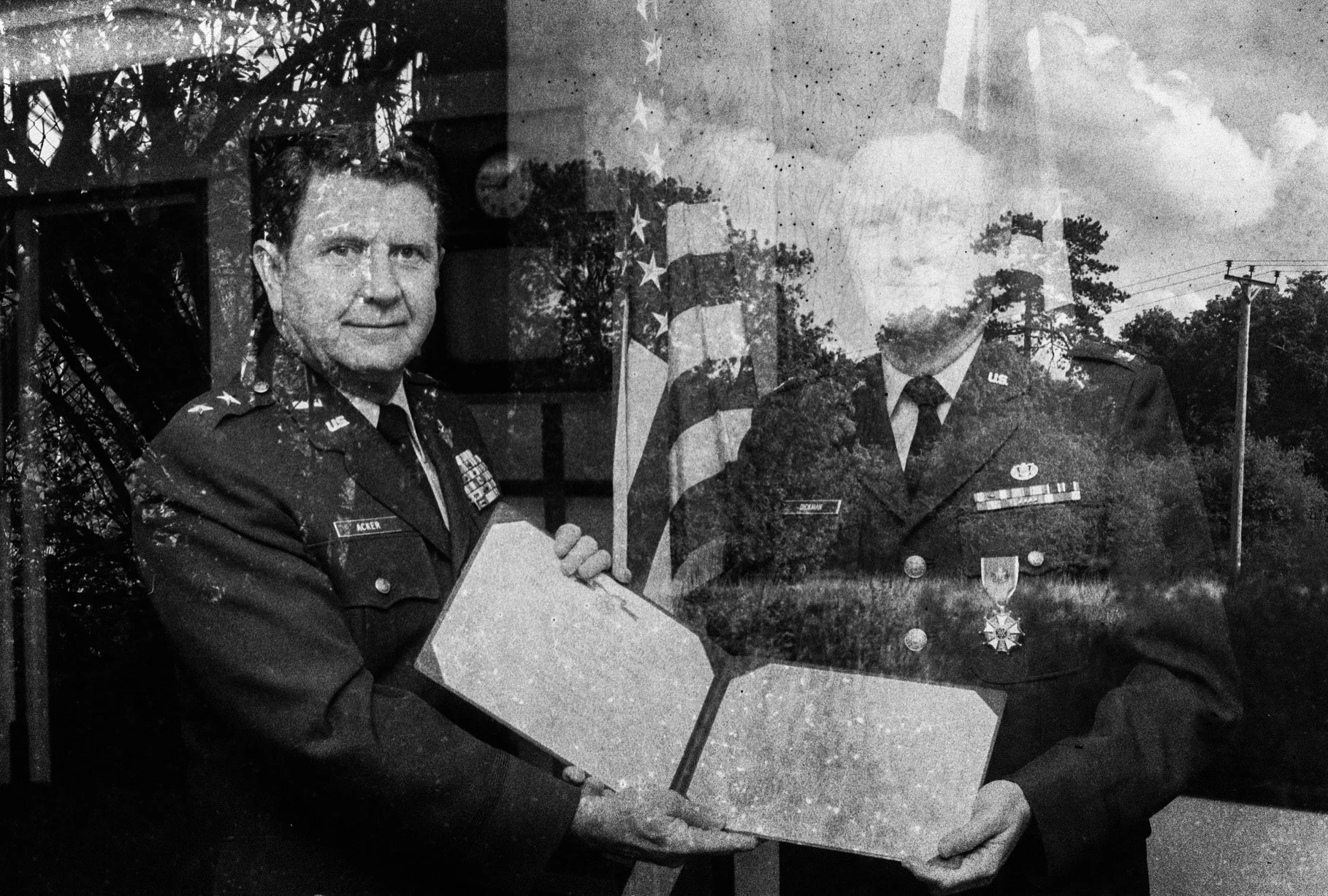 Black and white photo of two U.S. military officers in uniform holding a rectangular object, with an American flag and outdoor trees in the background.