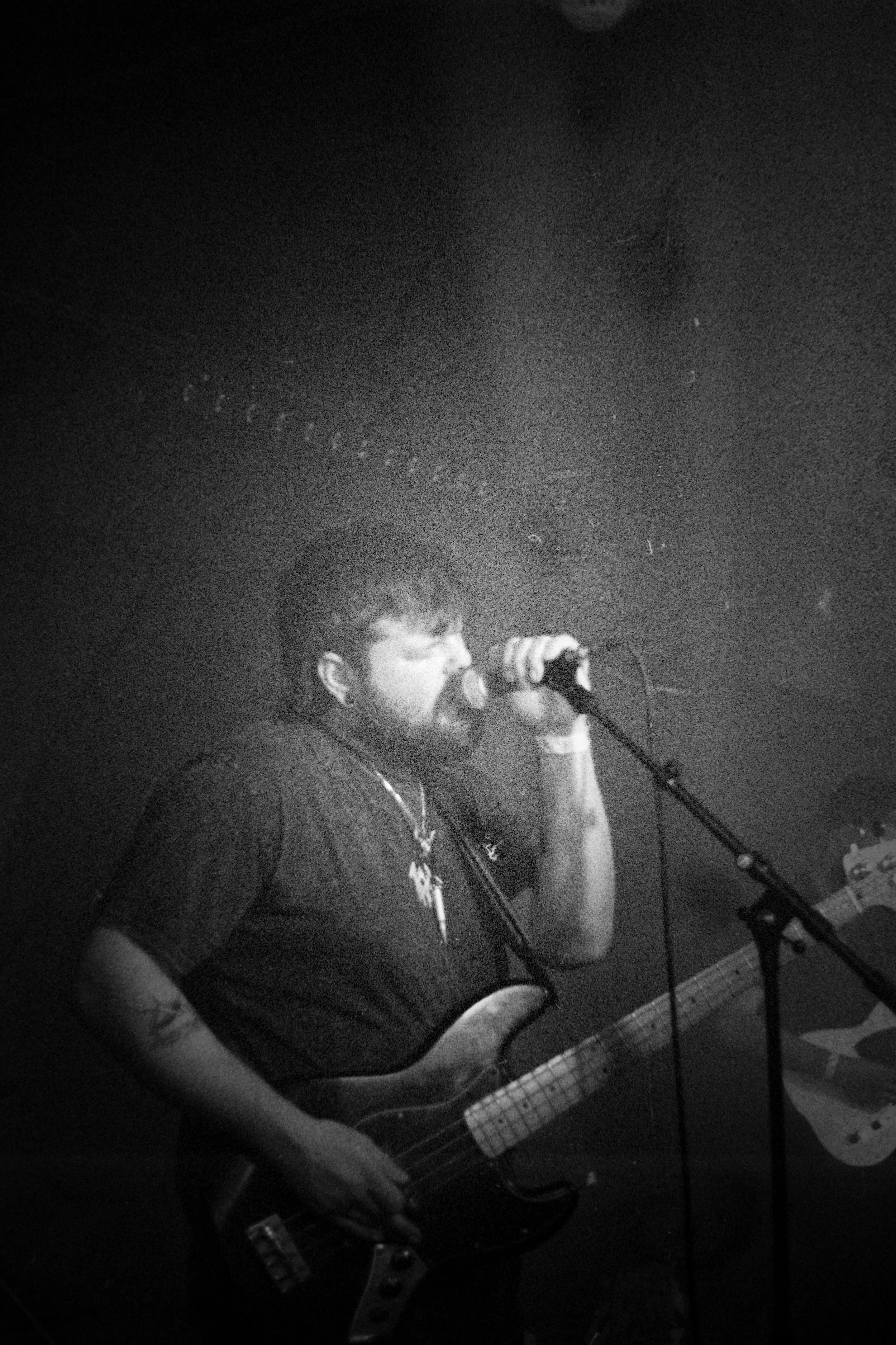 A man with a beard and tattoos is singing into a microphone while playing an electric guitar in a dark, smoky setting.