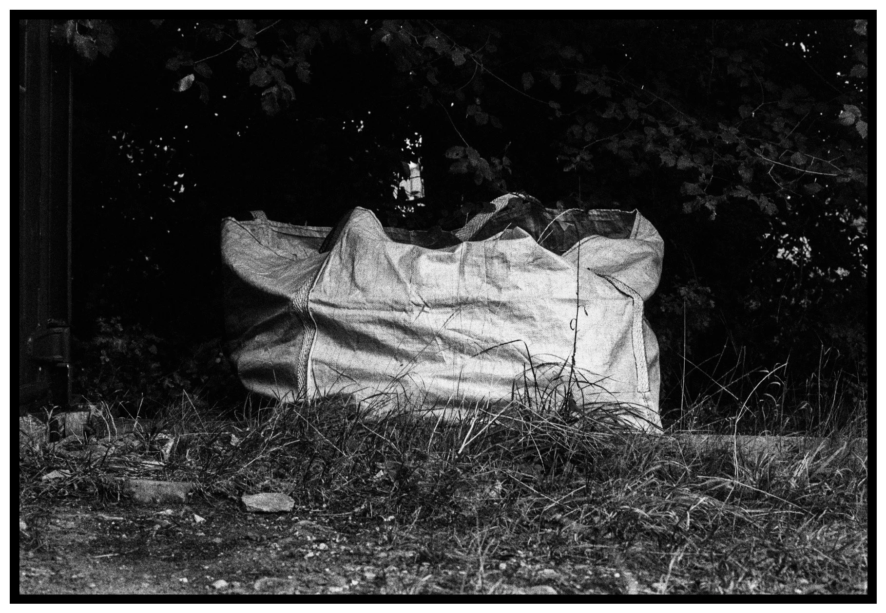 A large bag or tarp on the ground outdoors with grass and dirt in front and trees in the background.