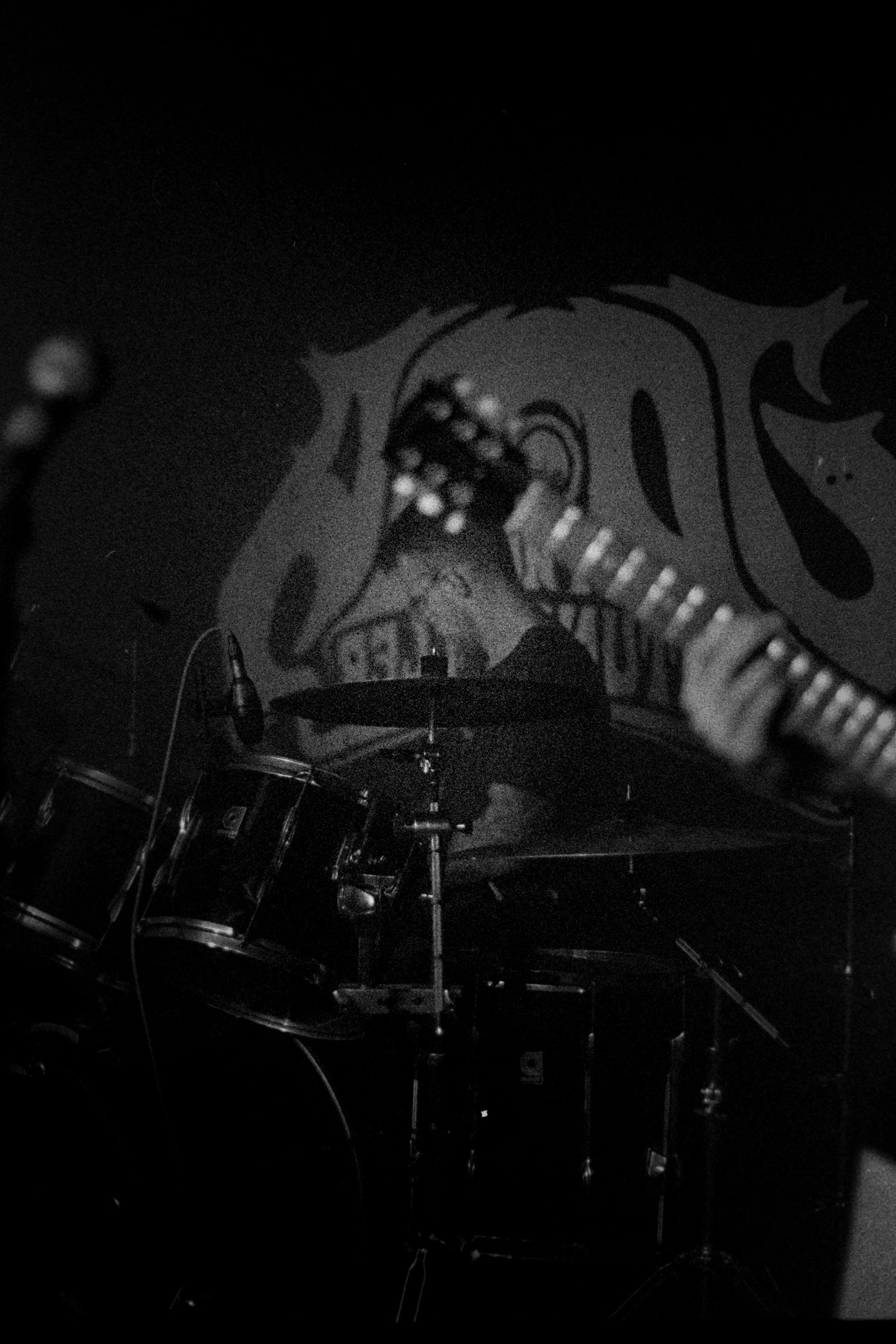 Black and white photo of a drum set on a stage with a guitar in the foreground and a wall decoration in the background.