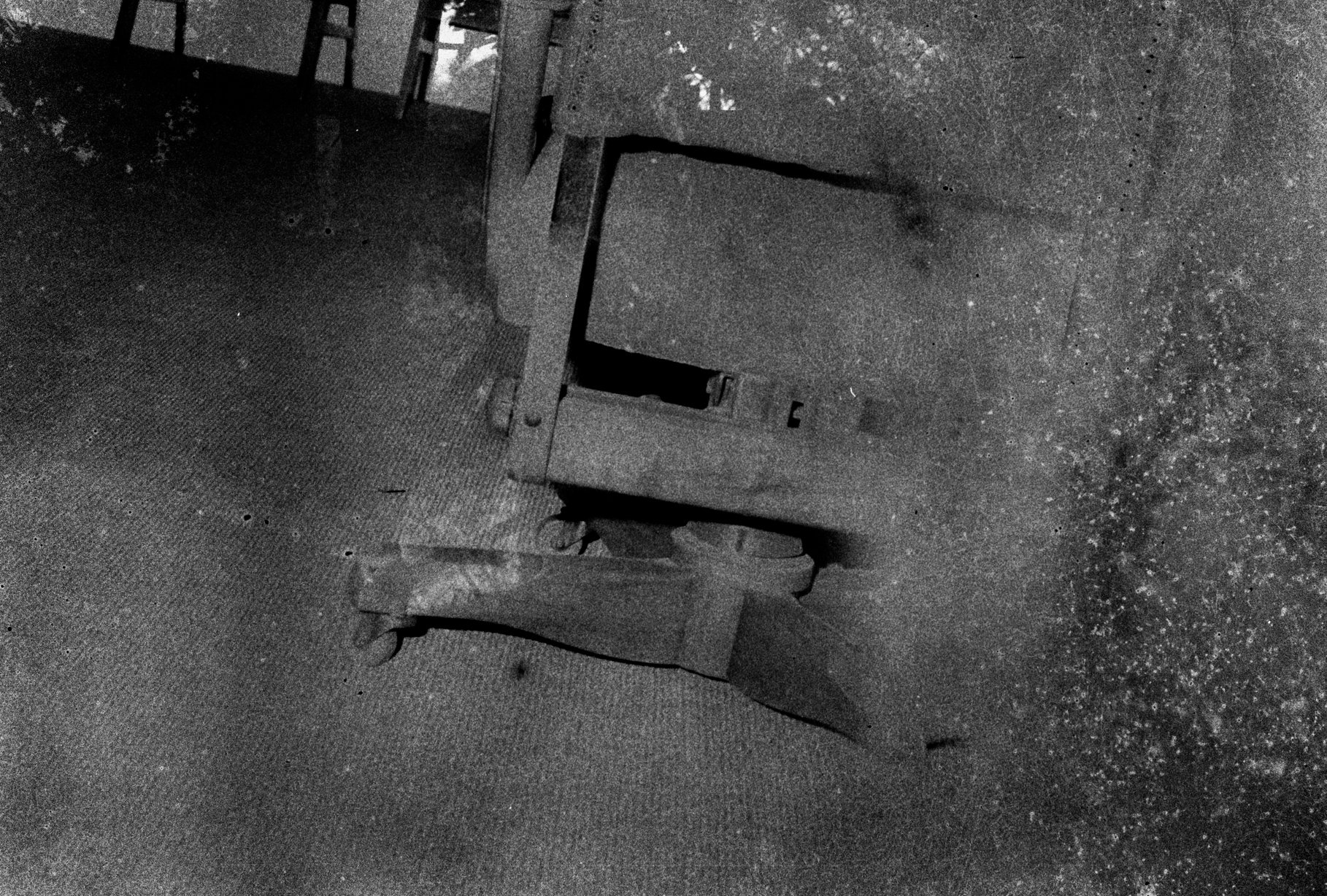 A black and white photo of a broken wooden wheelchair on a textured floor, with parts of a wooden table in the background.