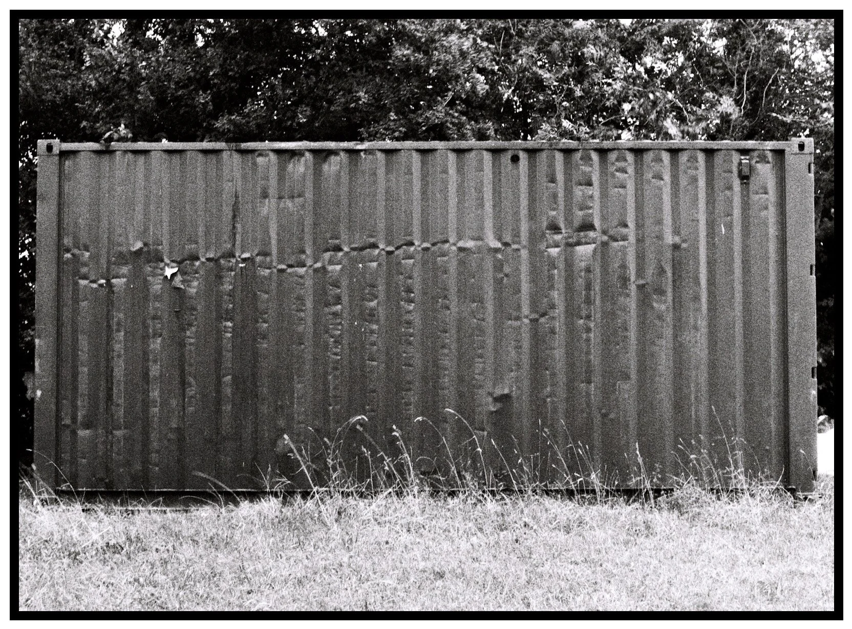 Black and white photo of a weathered metal shipping container on grassy ground, with trees in the background.