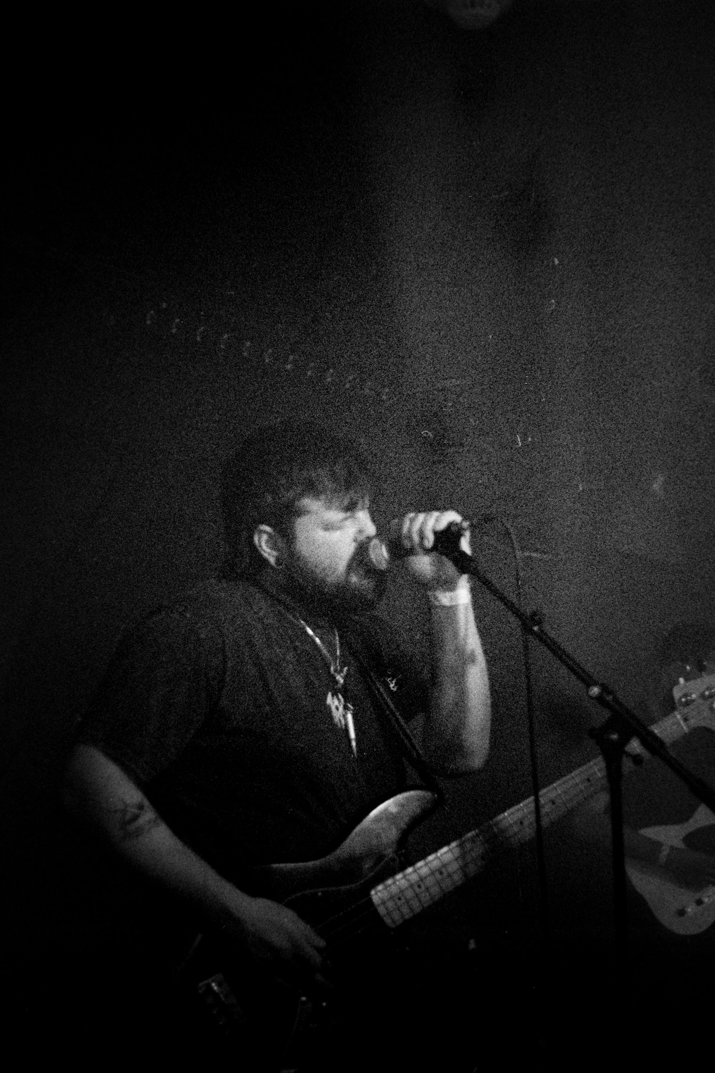 A man with a beard and tattoos singing into a microphone while playing an electric bass guitar in a dark setting