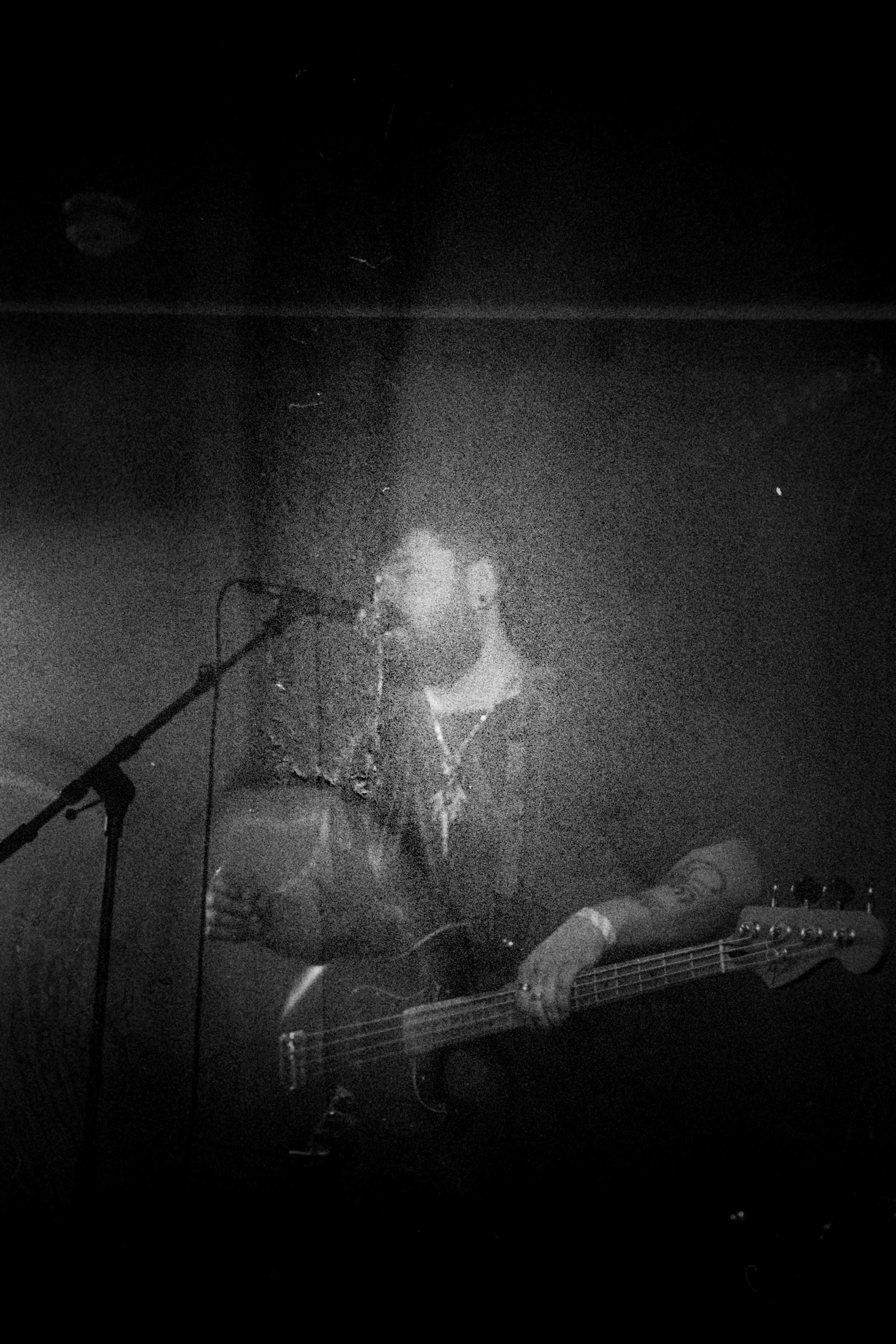 A musician singing into a microphone and playing an electric bass guitar, with a dark, smoky atmosphere.
