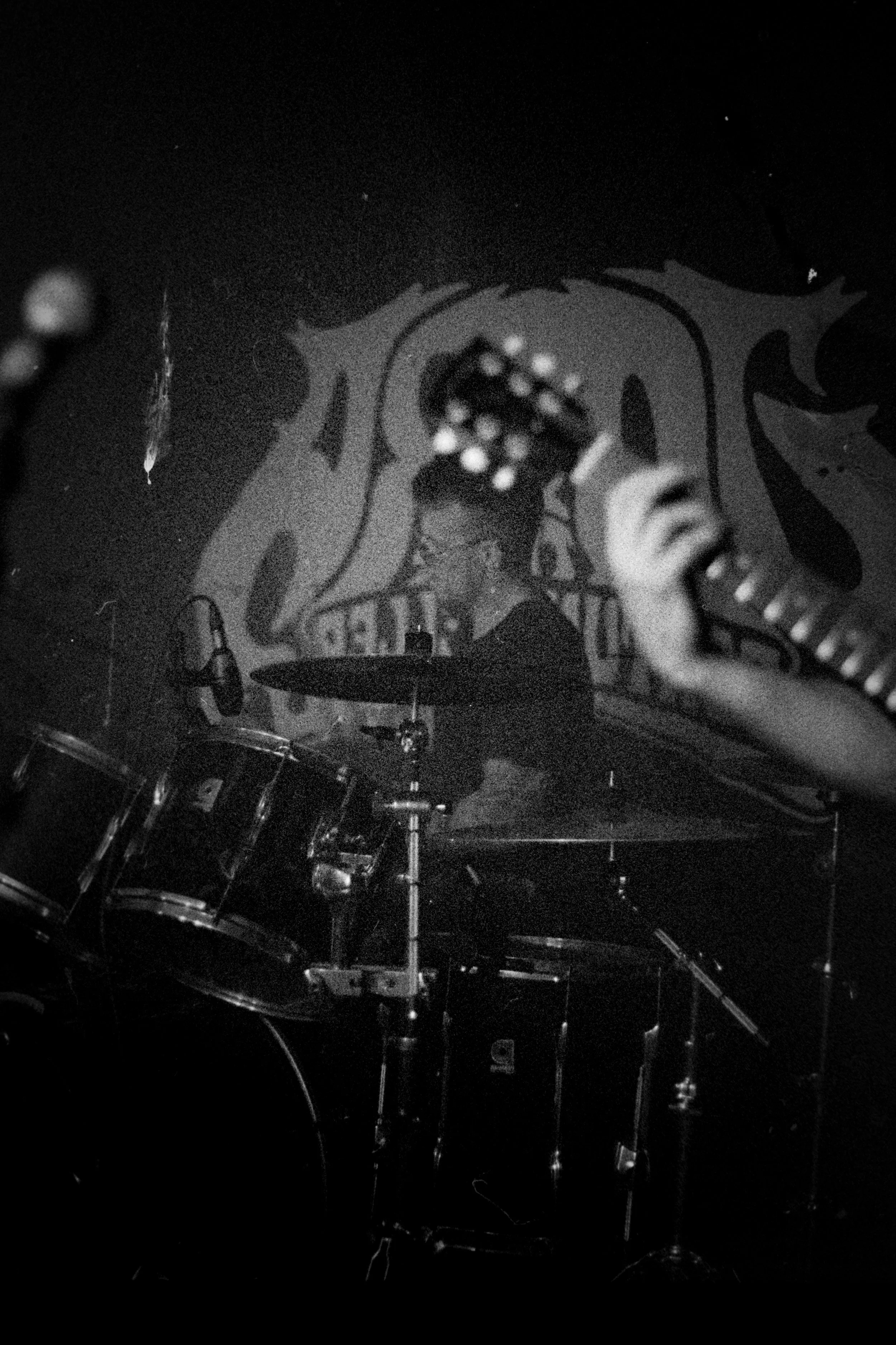 Black and white photo of a musician playing drums, with a mural or artwork in the background.