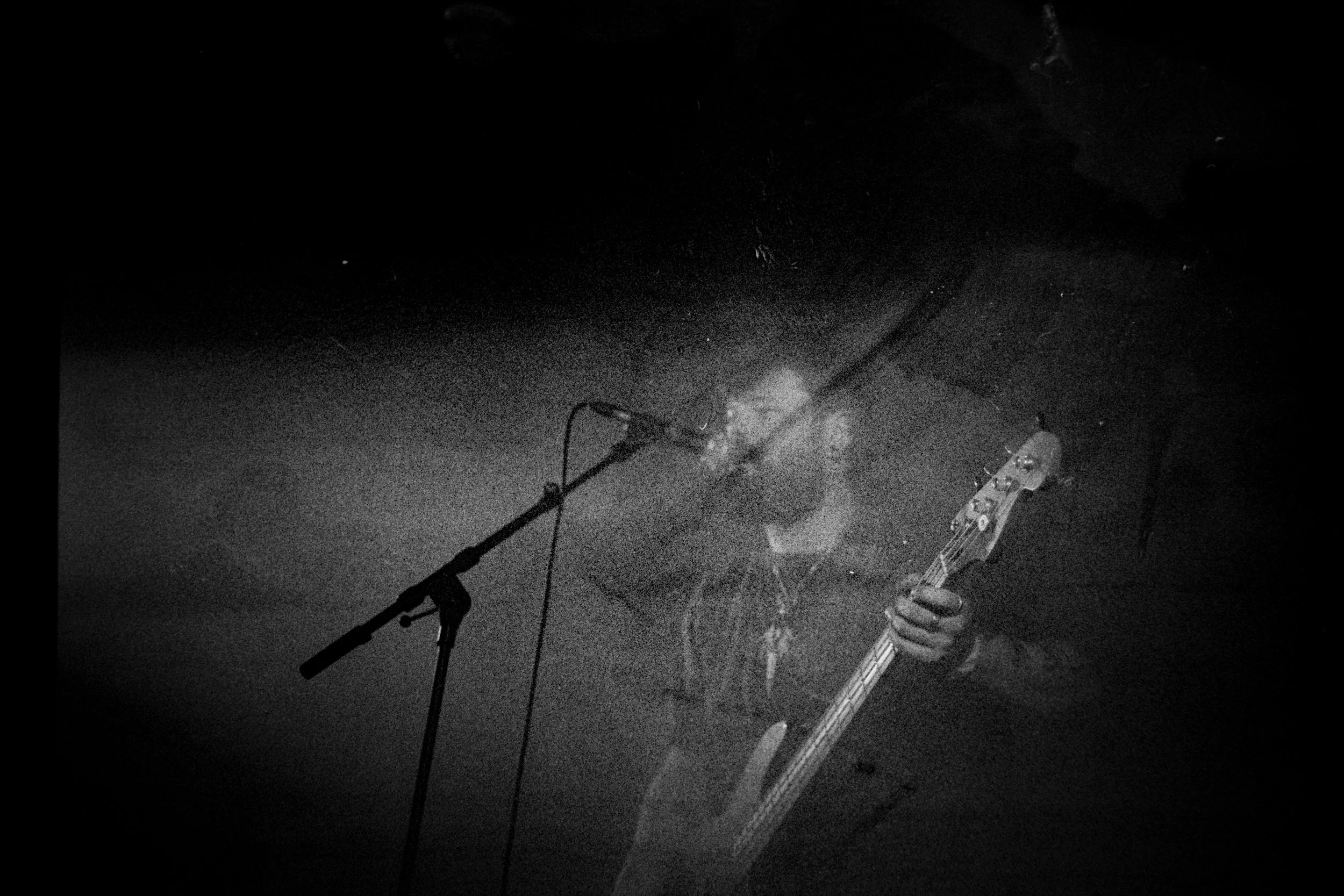 Black and white photo of a person playing an electric guitar and singing into a microphone on a dark stage.