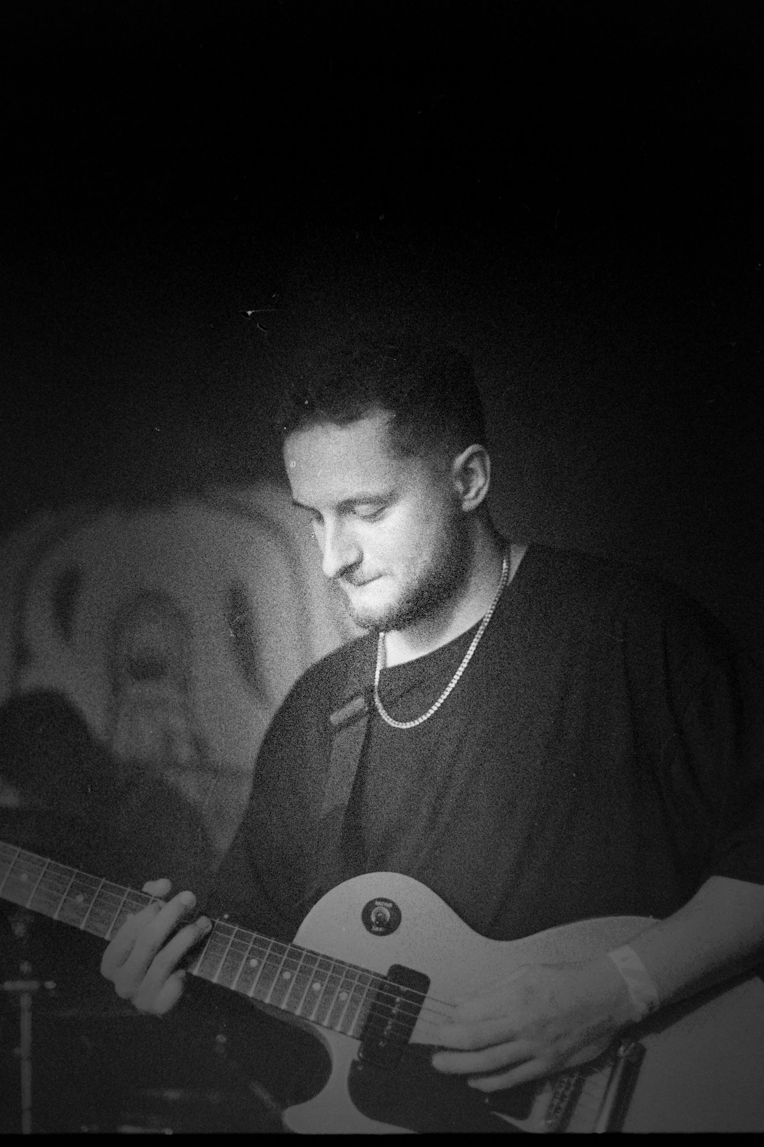 A young man with short hair playing an electric guitar in a dark setting, possibly a concert or rehearsal space.