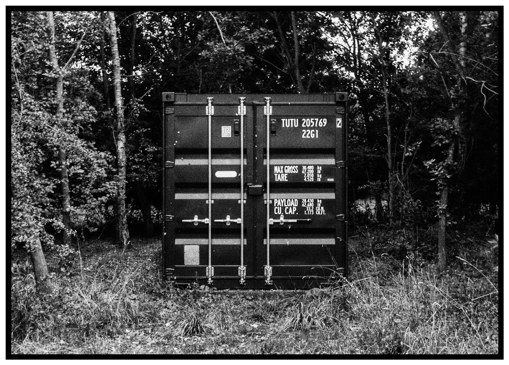 A black shipping container in a wooded area, surrounded by trees and grass.