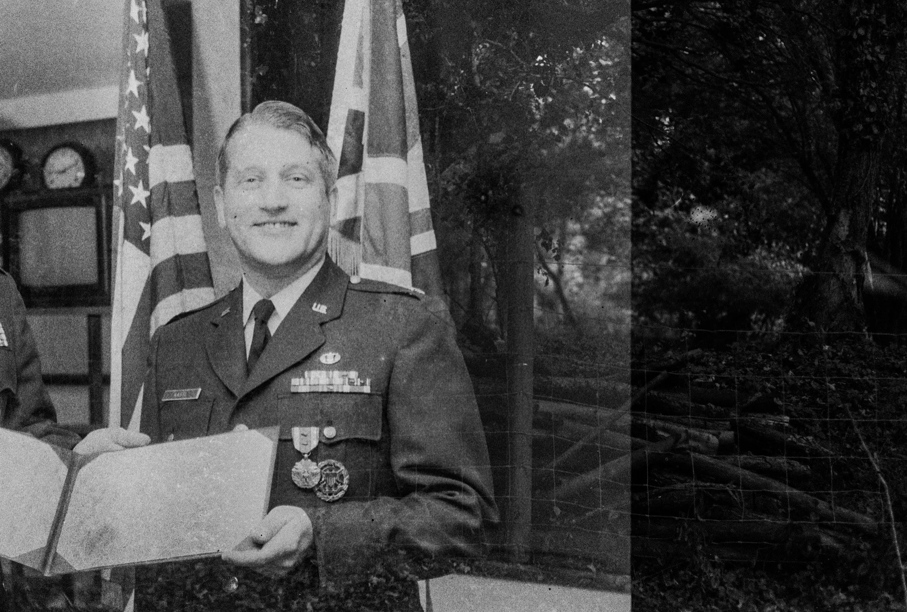 Black and white photograph of a soldier in uniform holding a document, standing in front of American flags and a Union Jack flag. Background shows trees and a wooded area.