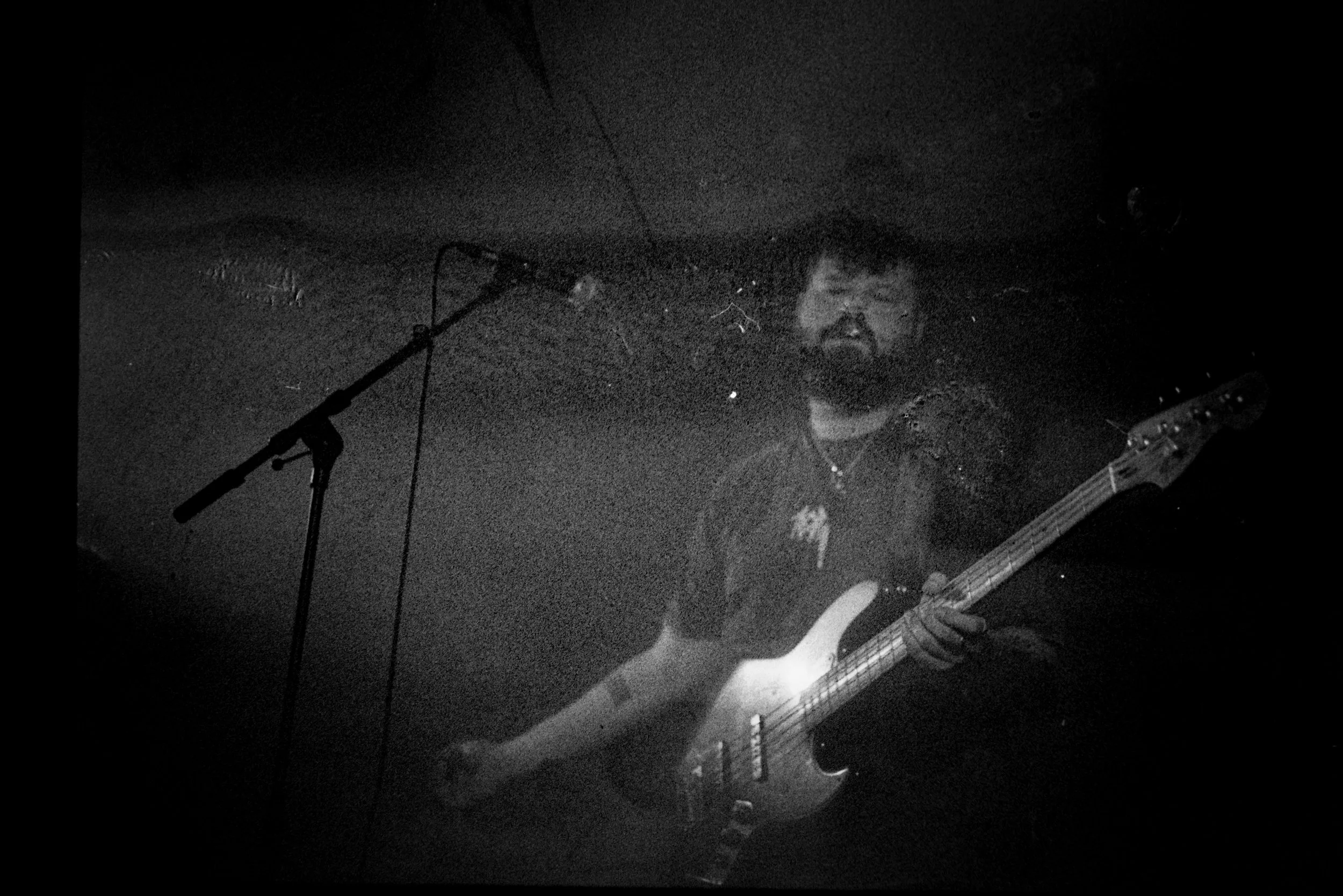 A man with a beard playing an electric bass guitar on stage, standing near a microphone in a dark setting.