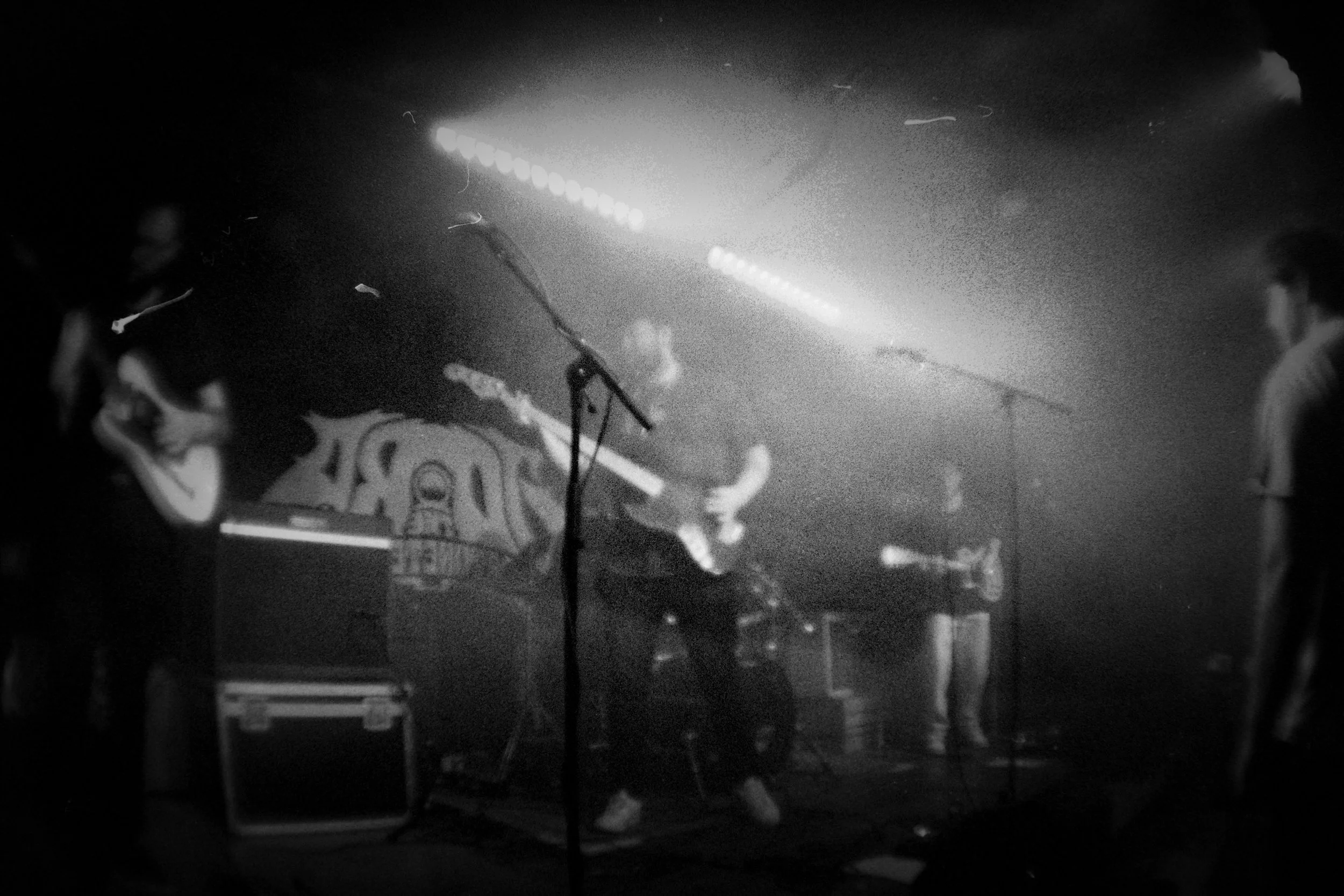A black and white photo of a band performing on stage with guitarists, a musician on keyboard, and another on bass, in a dimly lit venue with stage lights and a curtain backdrop.
