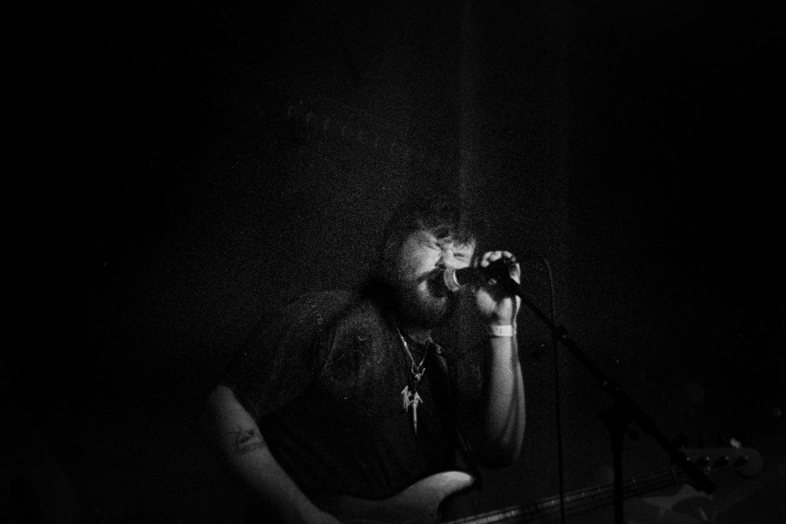 A musician with glasses, beard, and dark hair singing into a microphone while holding a guitar in a dimly lit setting.