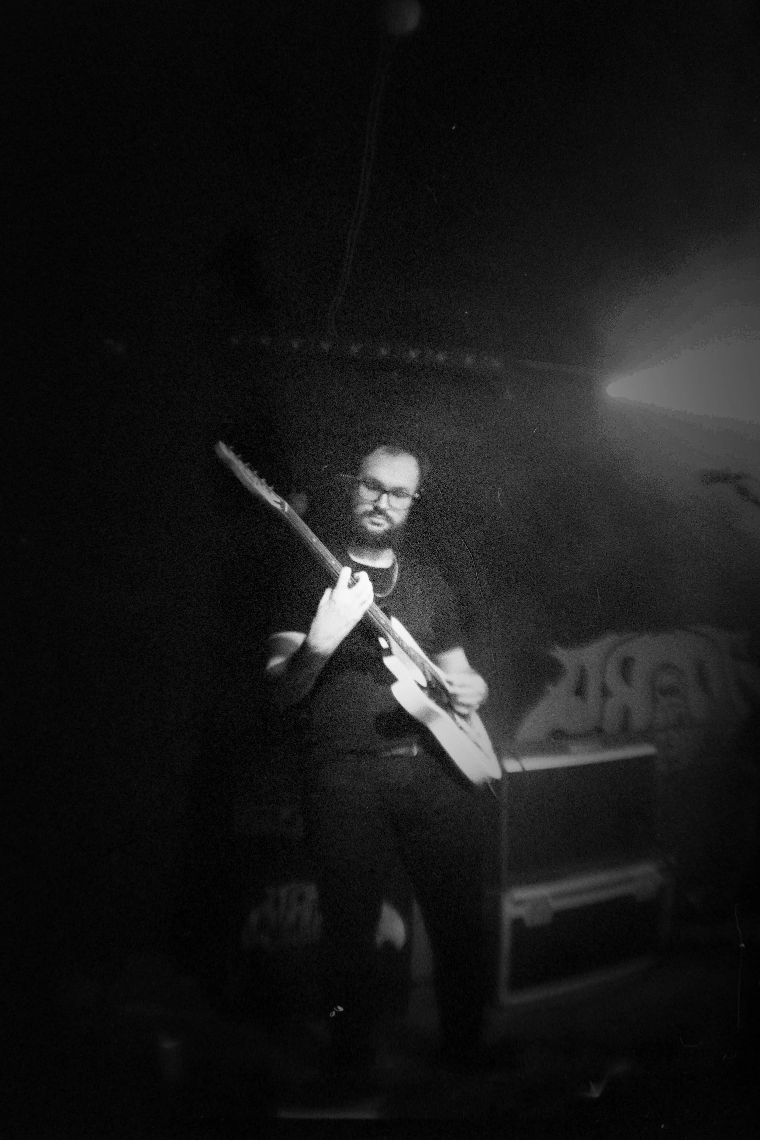 A man with glasses and a beard holding an electric guitar in a dark room with a bright light overhead.