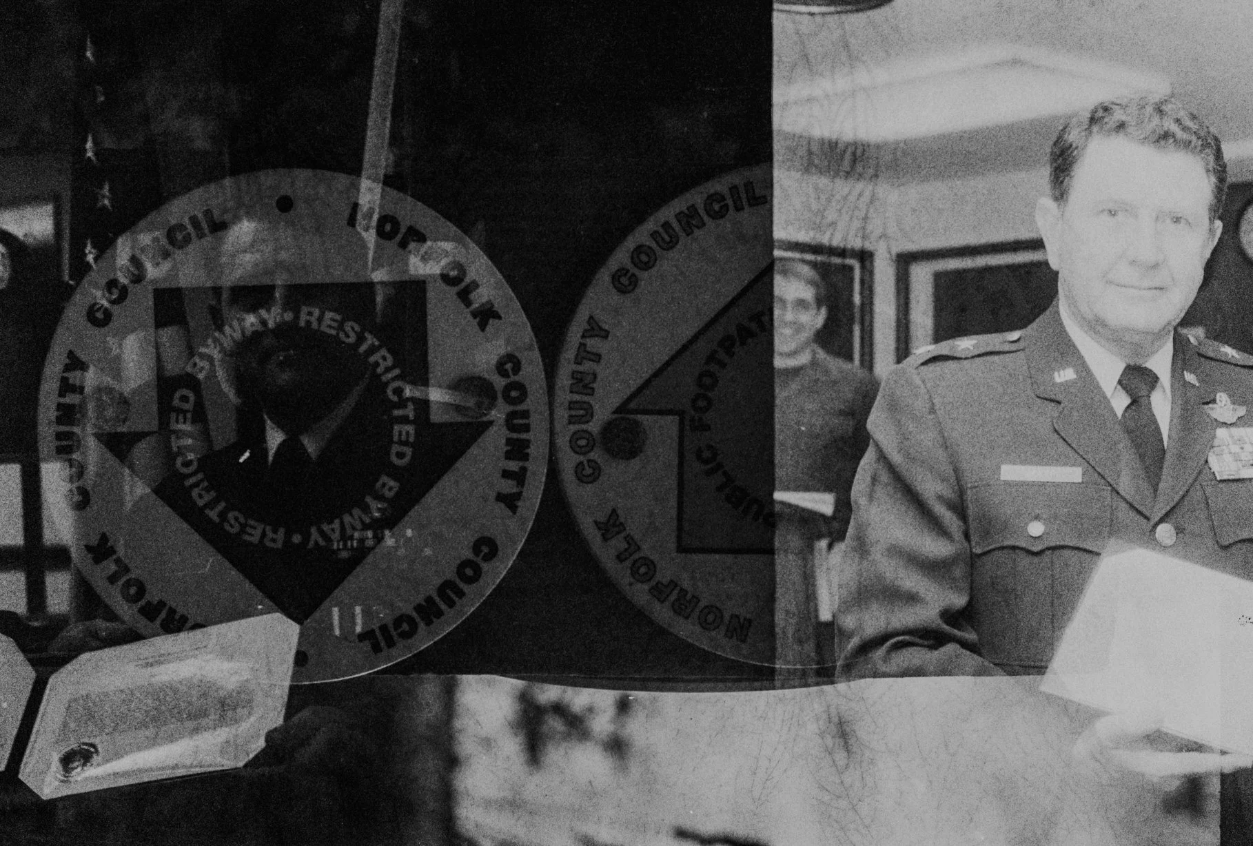 Black and white photo of a police officer in uniform with medals, holding a certificate, with a woman in uniform in the background.