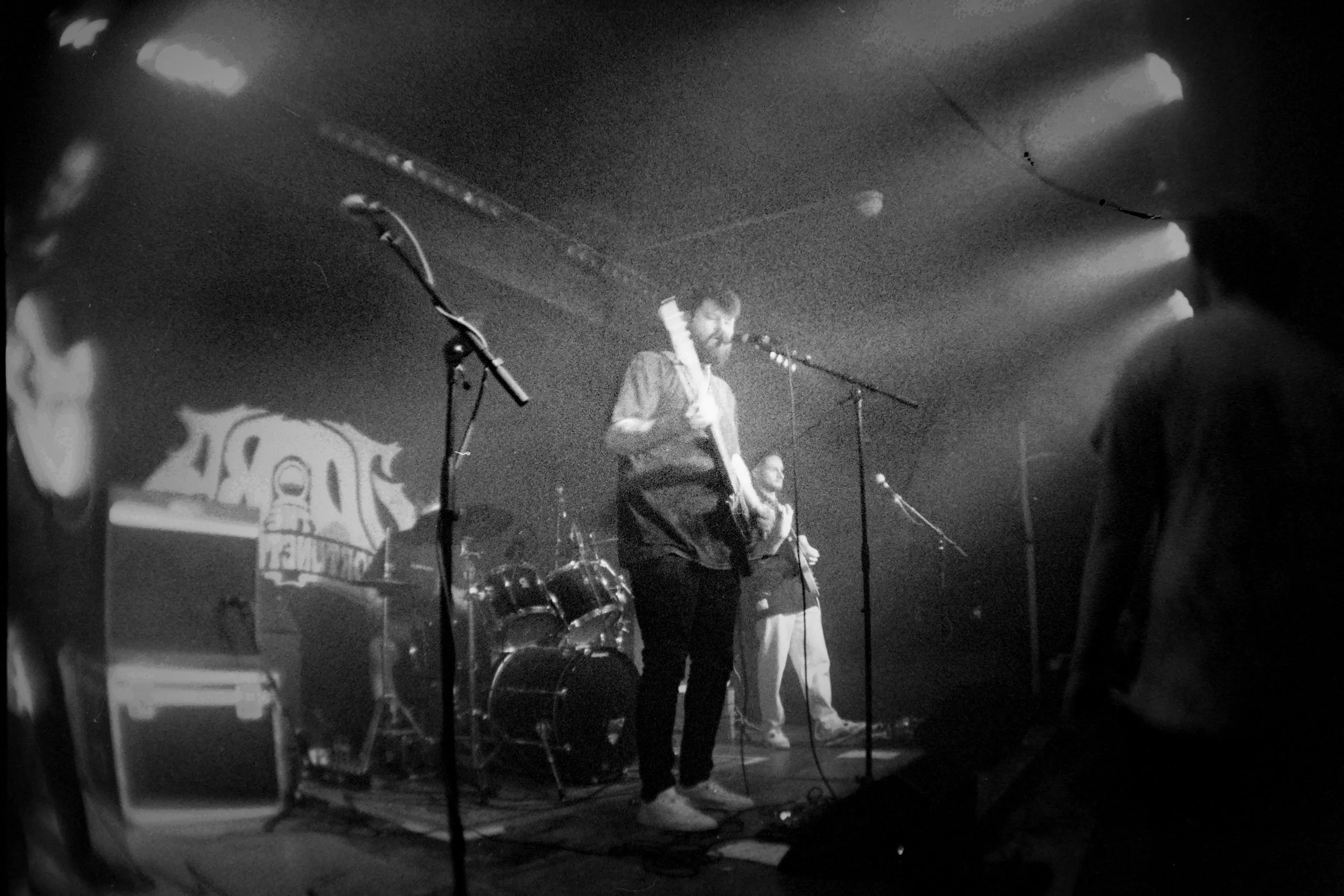 Black and white photo of a band performing on stage, with a drummer, a guitarist, and a singer in front of microphones.