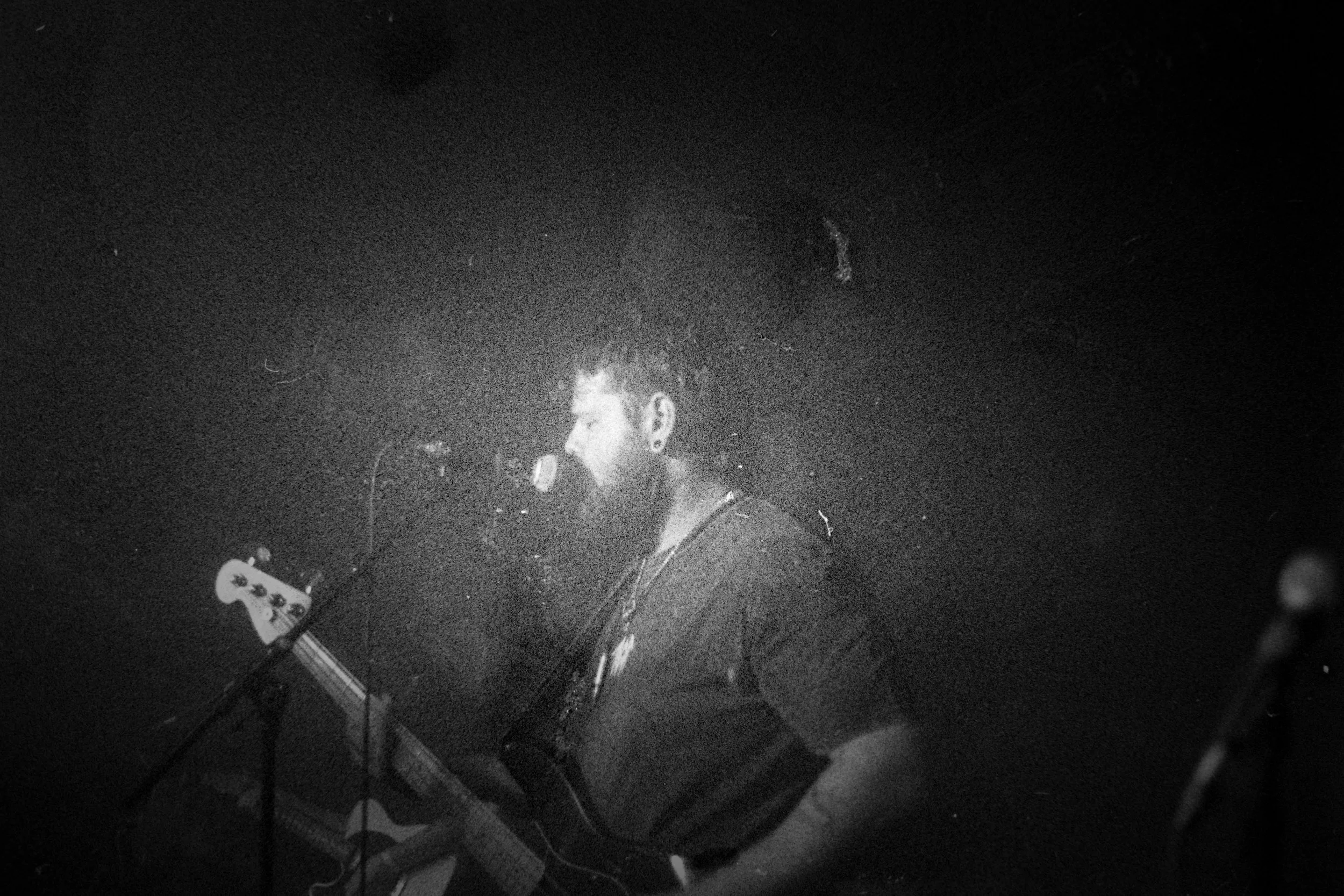 A black and white photo of a man singing into a microphone and playing an electric guitar on a dark stage.