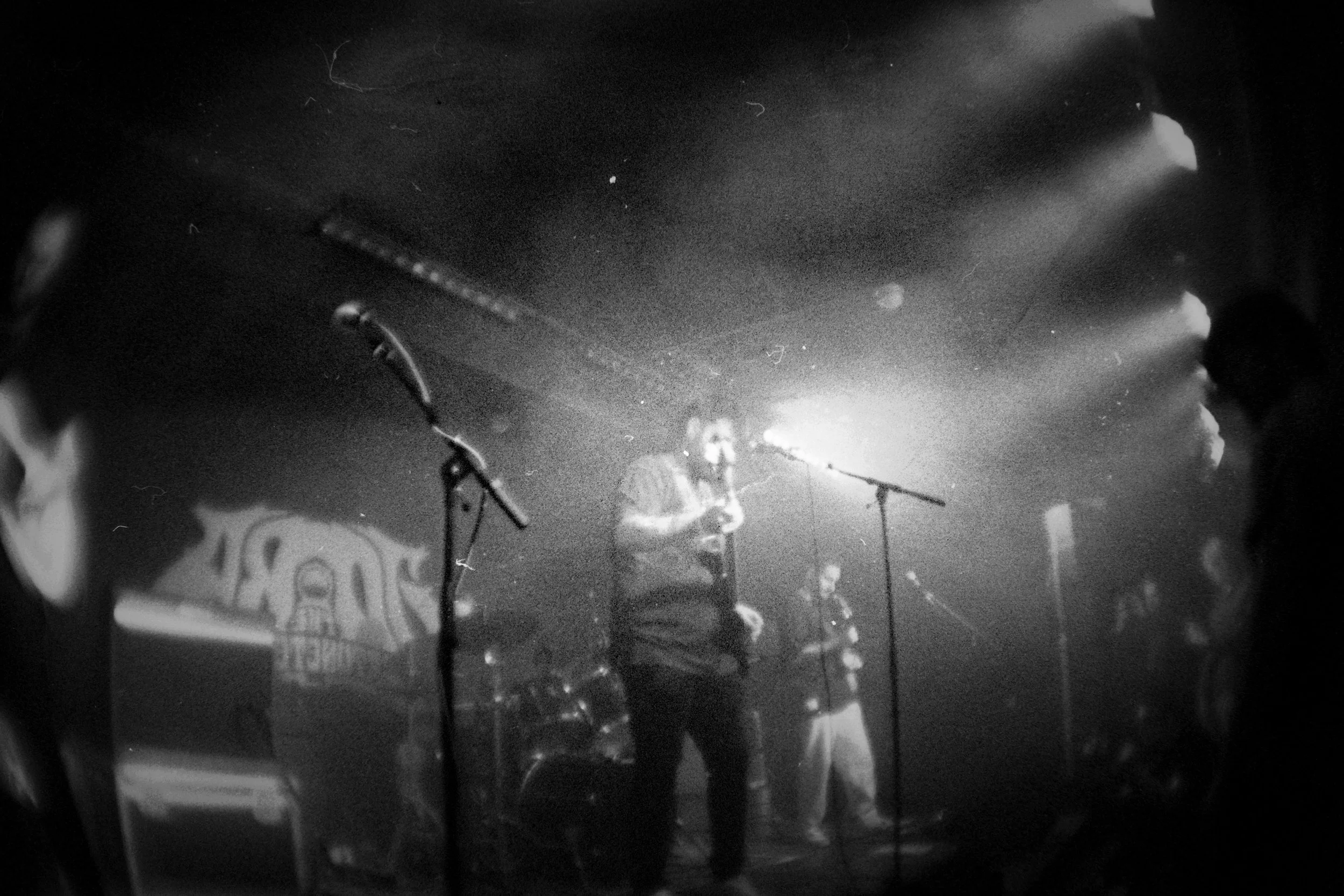 A black and white photo of a band performing on stage, with a man singing into a microphone, a guitarist, and other band members in the background, with stage lighting and equipment visible.