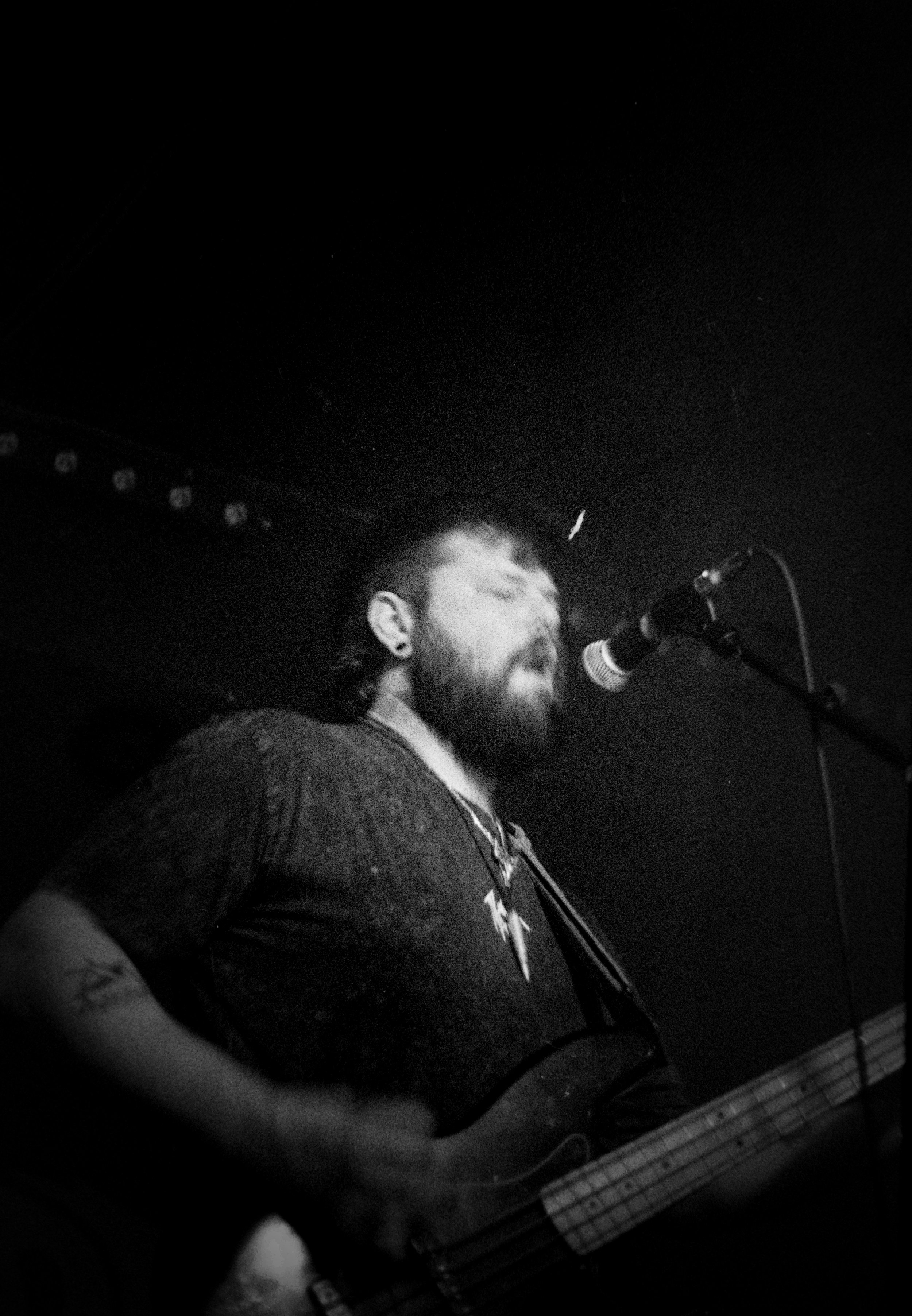 A black and white photo of a man singing into a microphone with his eyes closed, playing an electric bass guitar, on a dark stage.