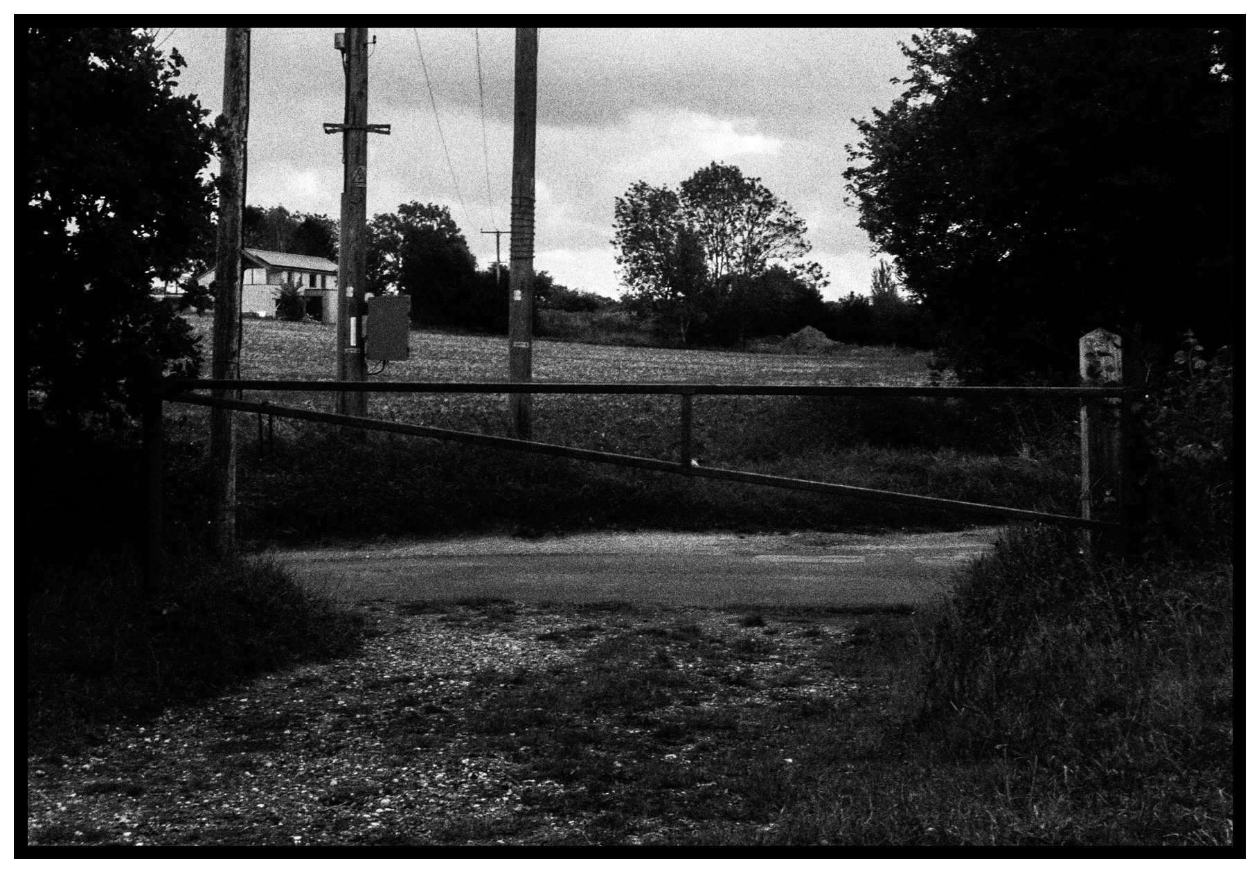 A black and white photo of a rural scene with a closed gate blocking a dirt and gravel road. Behind the gate, there are utility poles with power lines, trees, and a house on a slight hill.