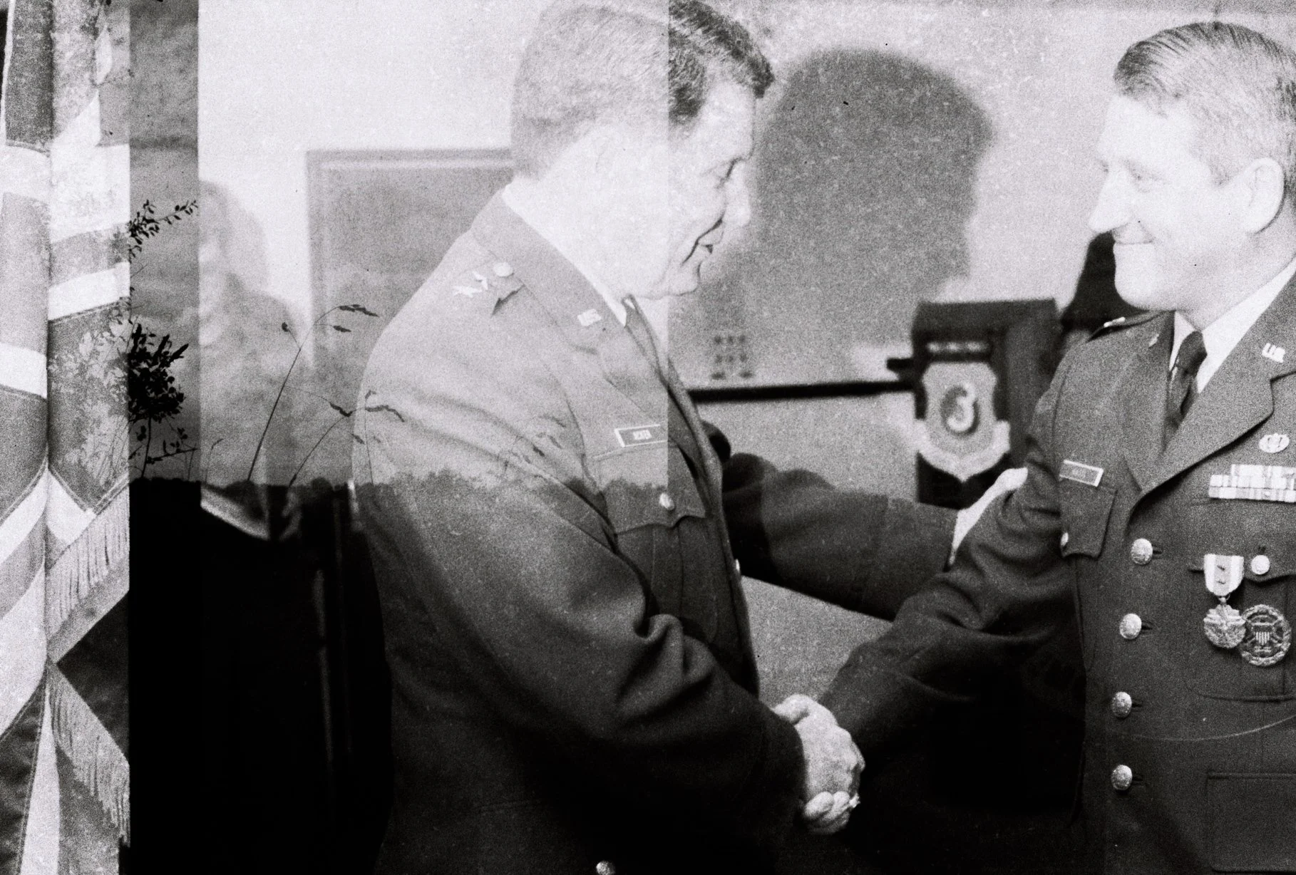 Two military officers shaking hands in an indoor setting. Both are dressed in uniform with medals, and there is a flag on the side.