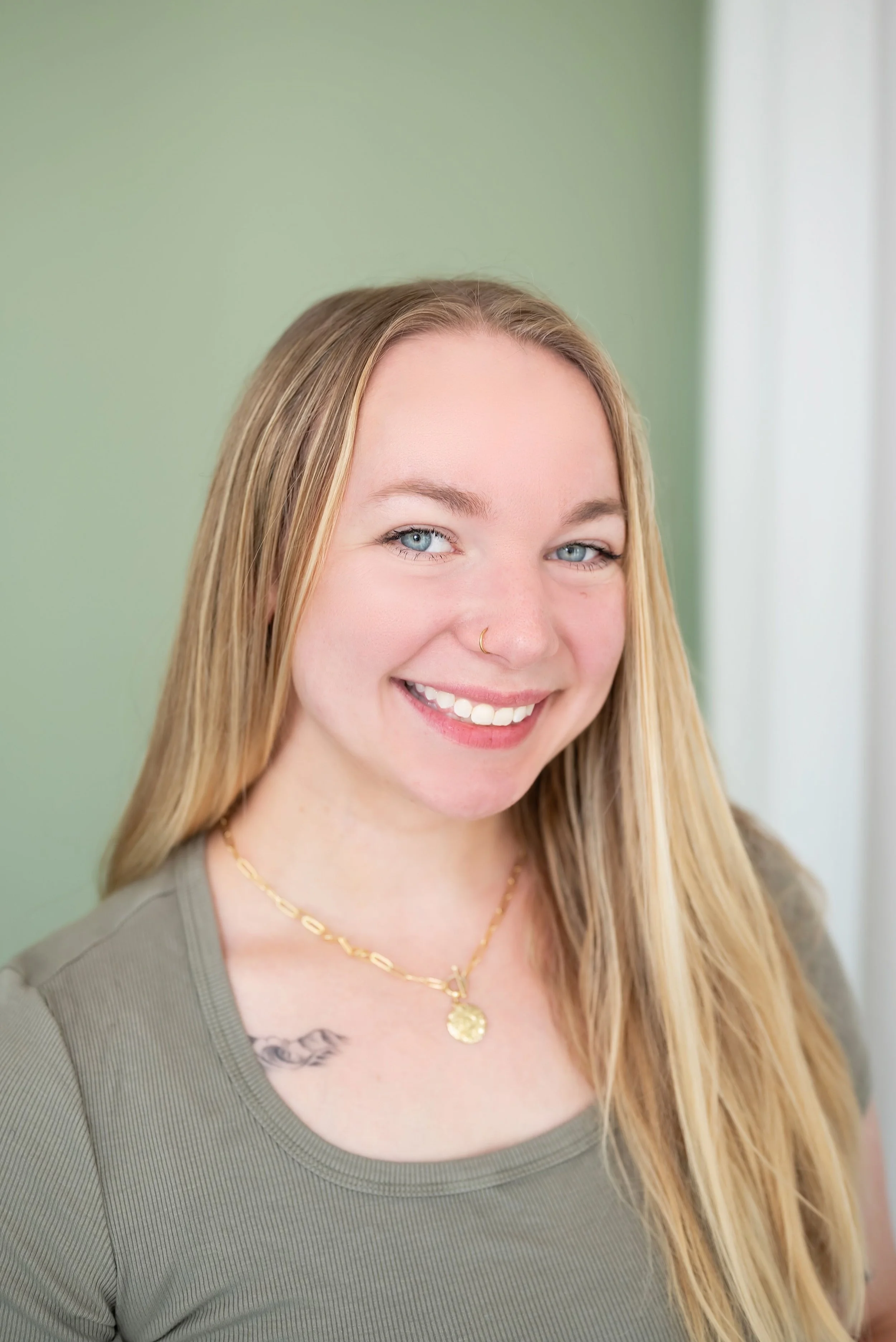Close-up of a young woman with long blonde hair, blue eyes, smiling, wearing gold jewelry and a green top, with a blurred green background.