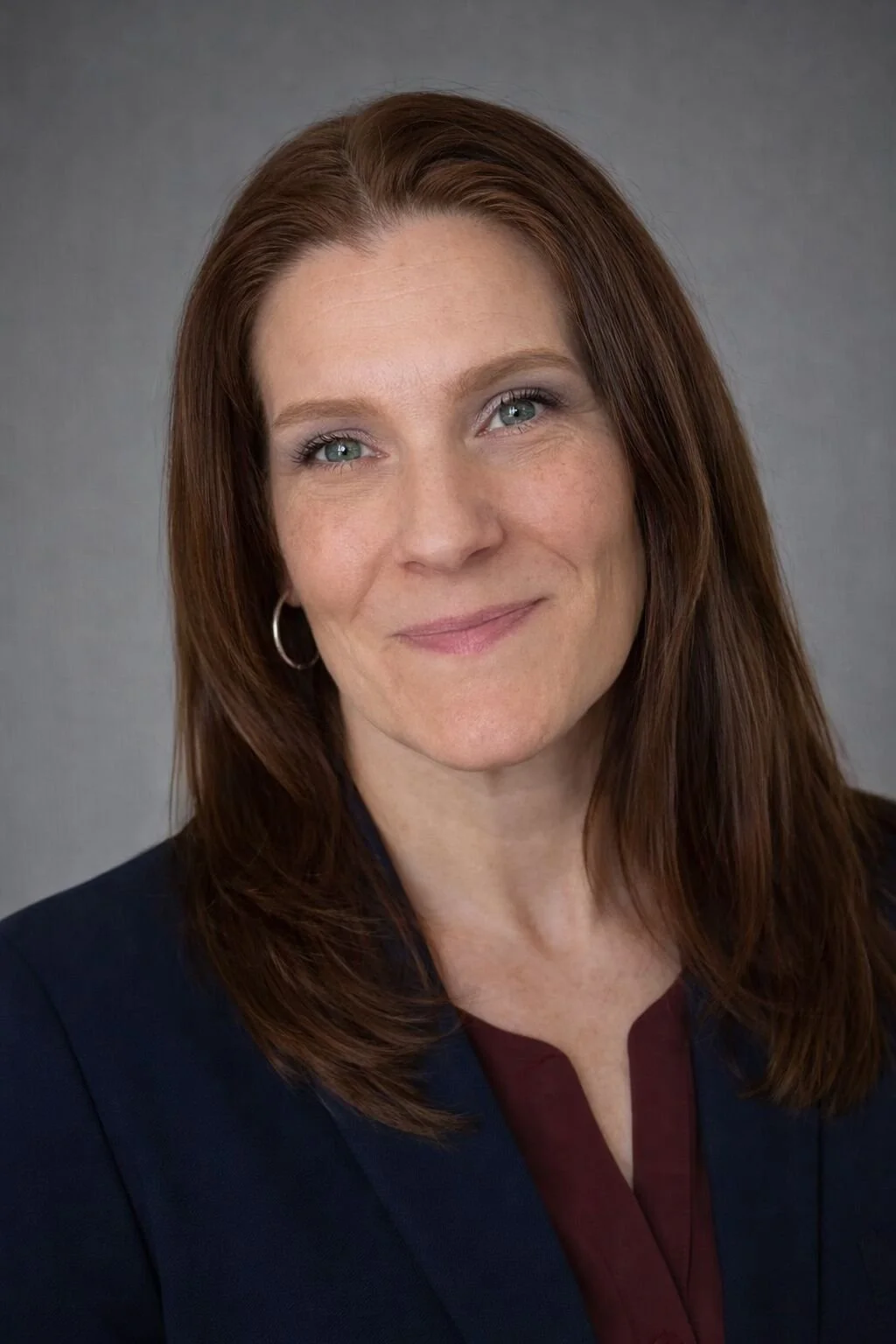 A woman with red hair, blue eyes, wearing hoop earrings, dressed in a dark blazer with a maroon top, smiling softly against a gray background.