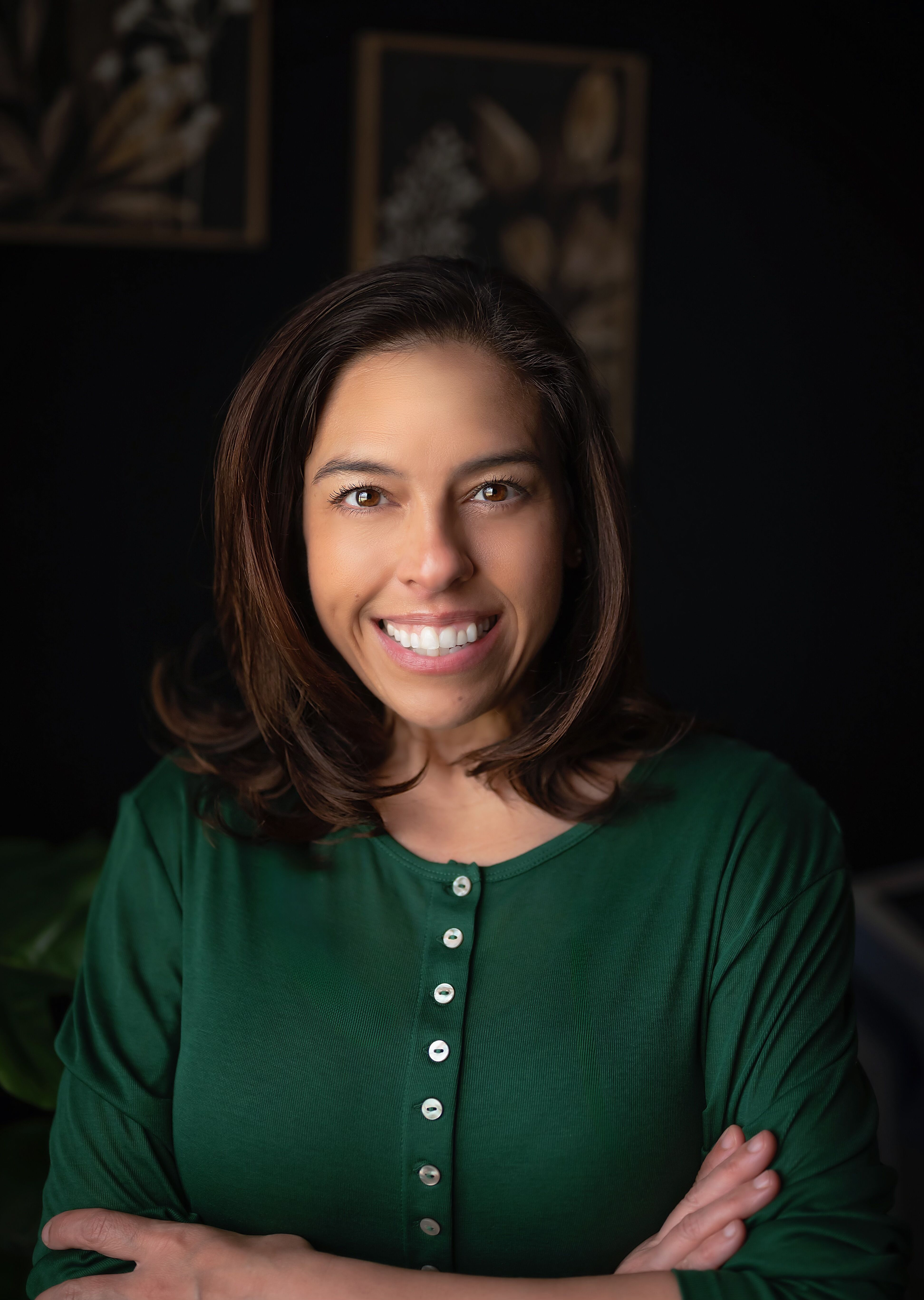 Close-up of a woman smiling, wearing a dark green top, with arms crossed, in front of dark background with blurred artwork.