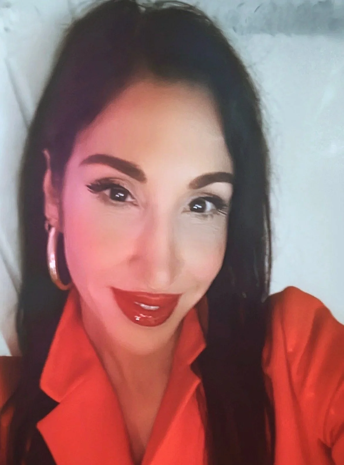A woman with long dark hair, wearing makeup, red lipstick, and hoop earrings, smiling in a selfie with a red top.