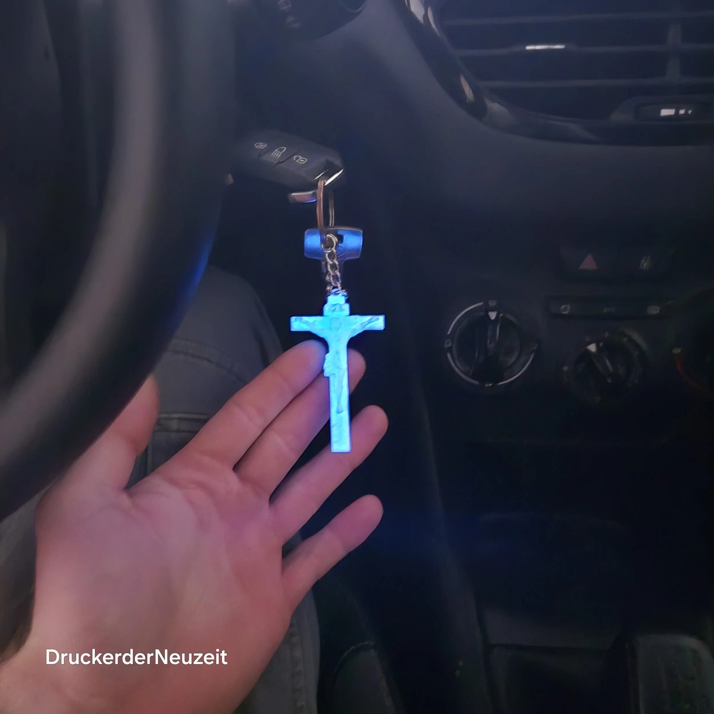 Hand hält einen leuchtenden blauen Kreuz-Anhänger im Auto, im Hintergrund Sicht auf die Armaturen und das Lenkrad.