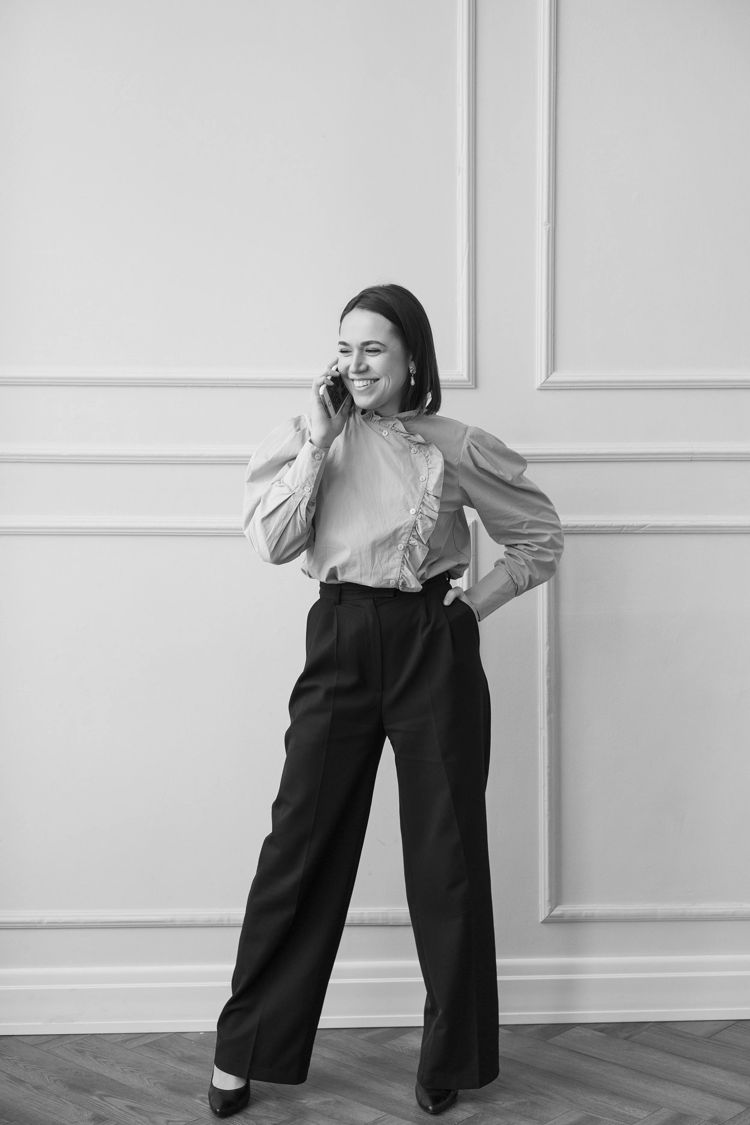 Une femme souriante parlant au téléphone, habillée d'une chemise à manches bouffantes et d'un pantalon large, debout dans une pièce élégante.