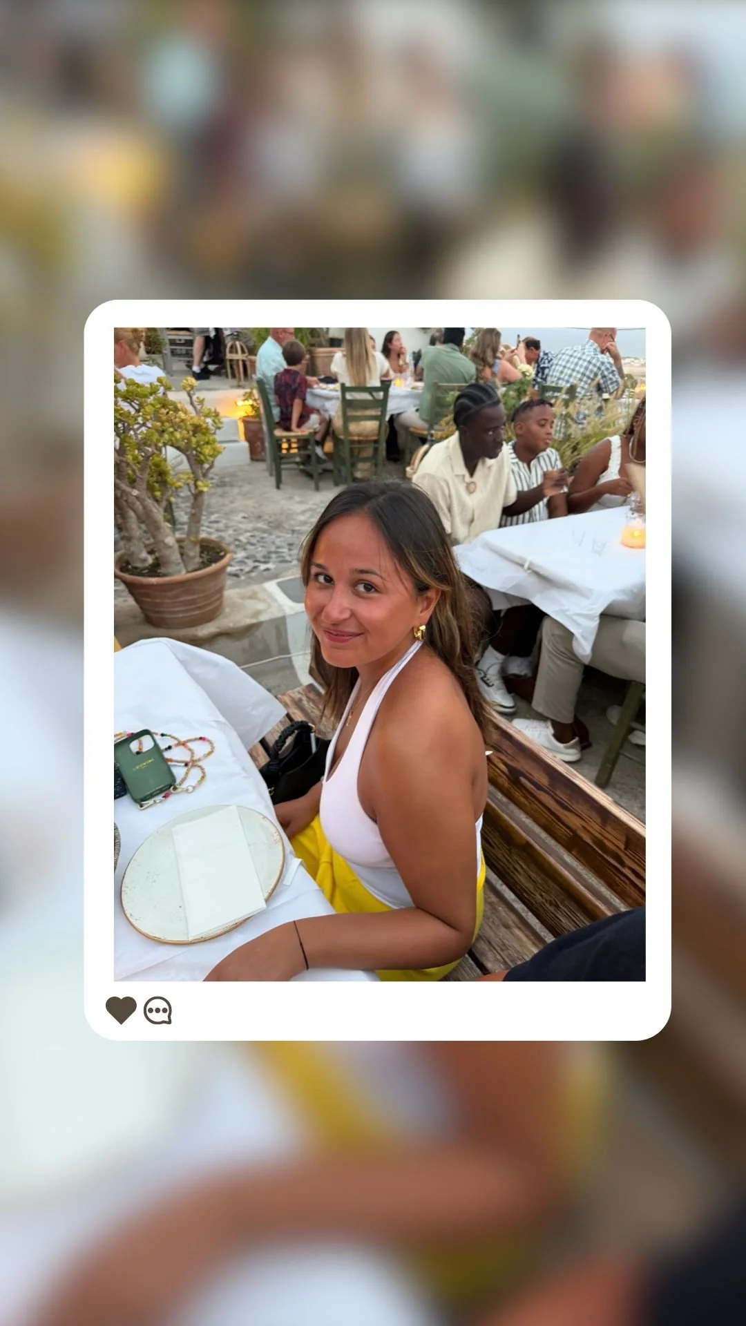 Une femme assise à une table dans un restaurant en plein air, entourée d'autres convives, avec une plante en pot à côté.