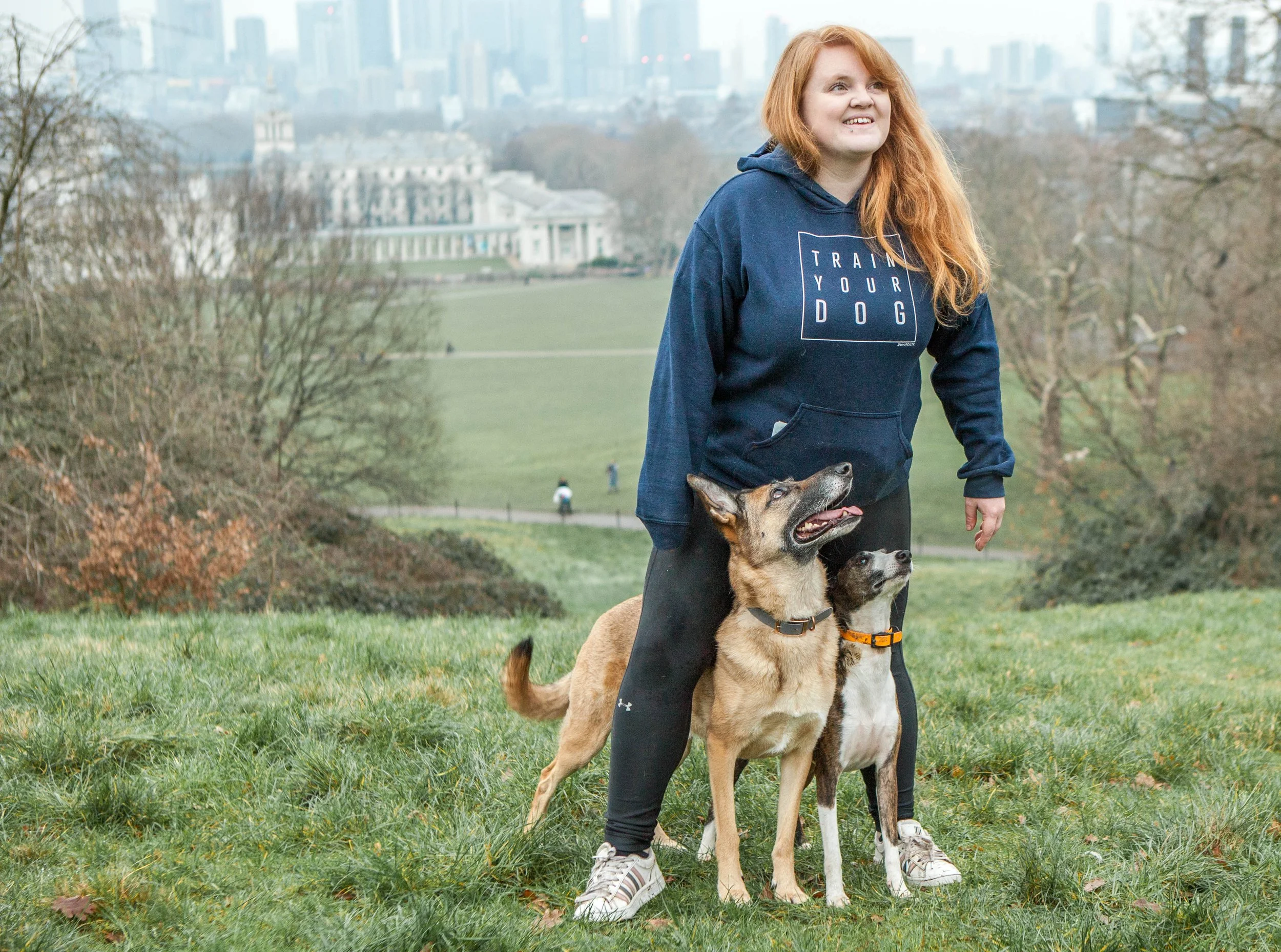 A girl with long red hair wearing a blue hoodie with the words 'Train Your Dog' on it, standing in a park with two dogs at her feet, one tan and black, the other black and white. The background shows a city skyline and trees.