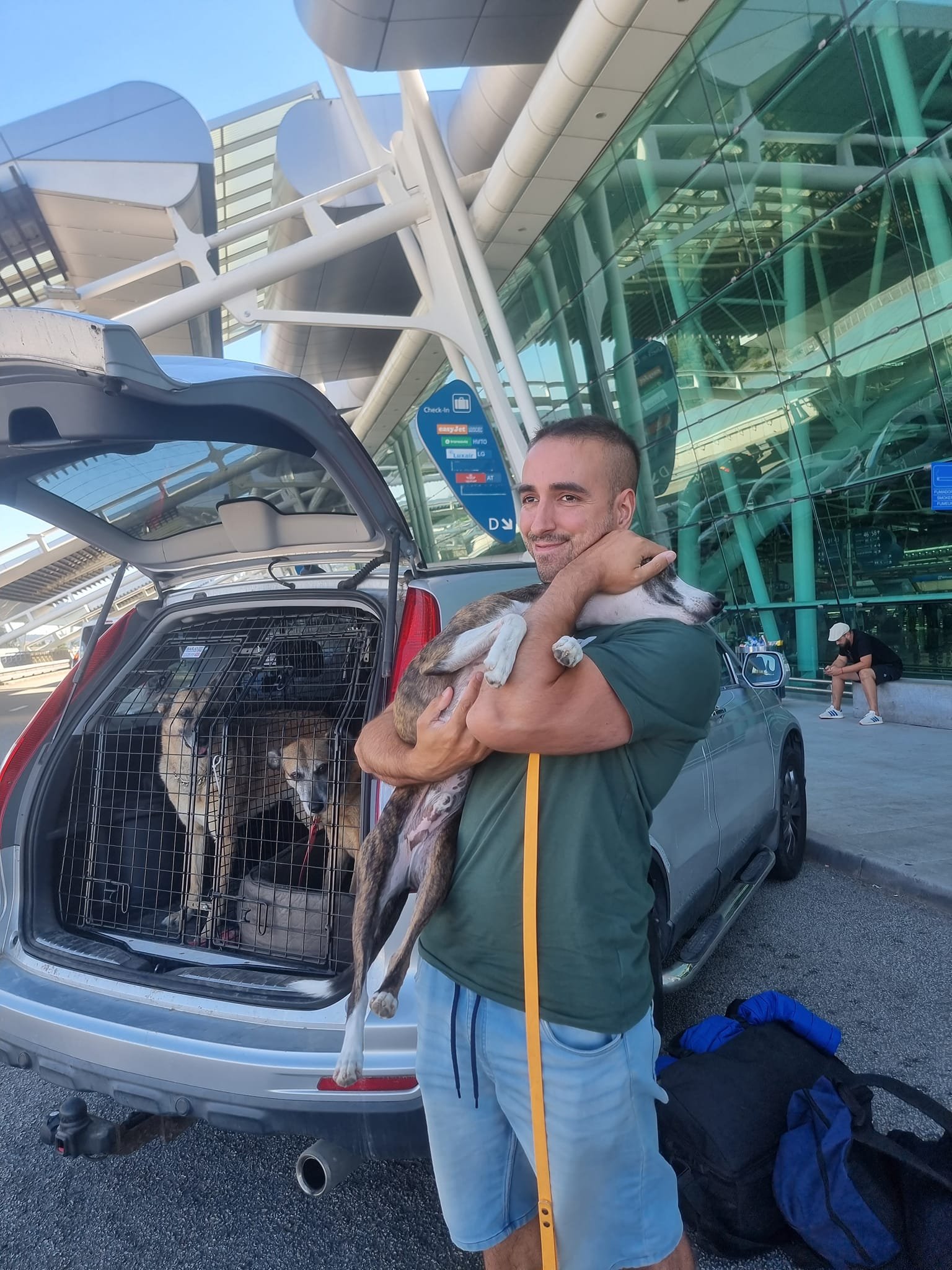 A man holding a dog outside an airport terminal with two dogs in the car's pet crate.