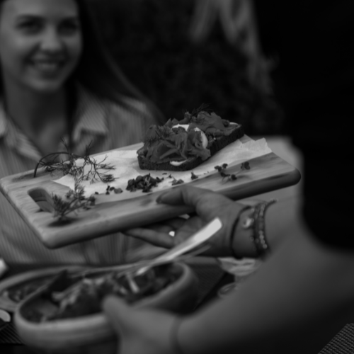 Black and white photo of a person serving plated food on a wooden board while diners sit at a table in the background.
