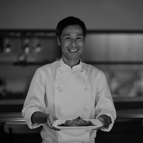 Smiling chef in a white uniform presenting a plated dish inside a modern restaurant kitchen.