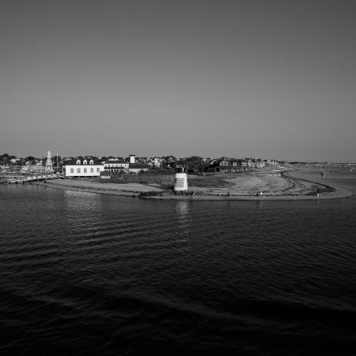 Black and white aerial photo of a coastal peninsula with a small lighthouse, shoreline homes, and calm water surrounding it.