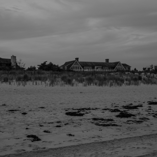 Black and white photo of a sandy beach with dune grass and large coastal homes under an overcast sky.