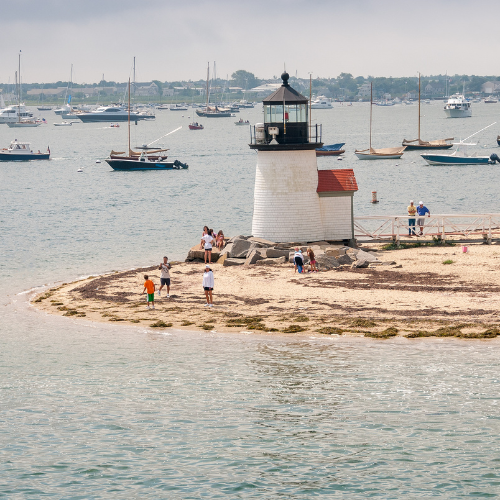 Small coastal lighthouse on a sandy shoreline with people nearby and boats anchored across the harbor.