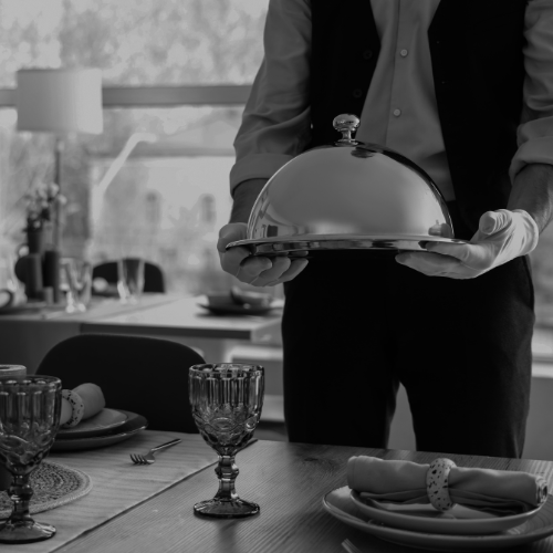 Black and white photo of a server holding a covered silver platter above a formally set dining table indoors.