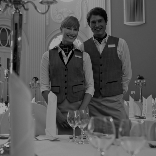 Black and white photo of two hospitality staff members standing in an elegant dining room with formally set tables.