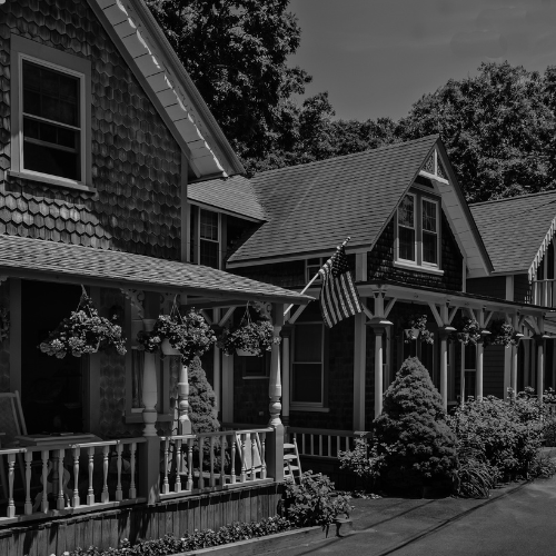 Black and white photo of charming cottage-style homes with shingled siding, front porches, hanging flower baskets, and an American flag along a quiet lane.