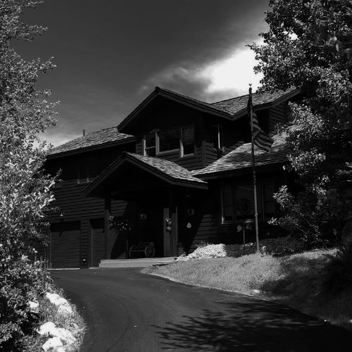 Black and white image of a large mountain lodge-style home with an American flag in a wooded residential area.